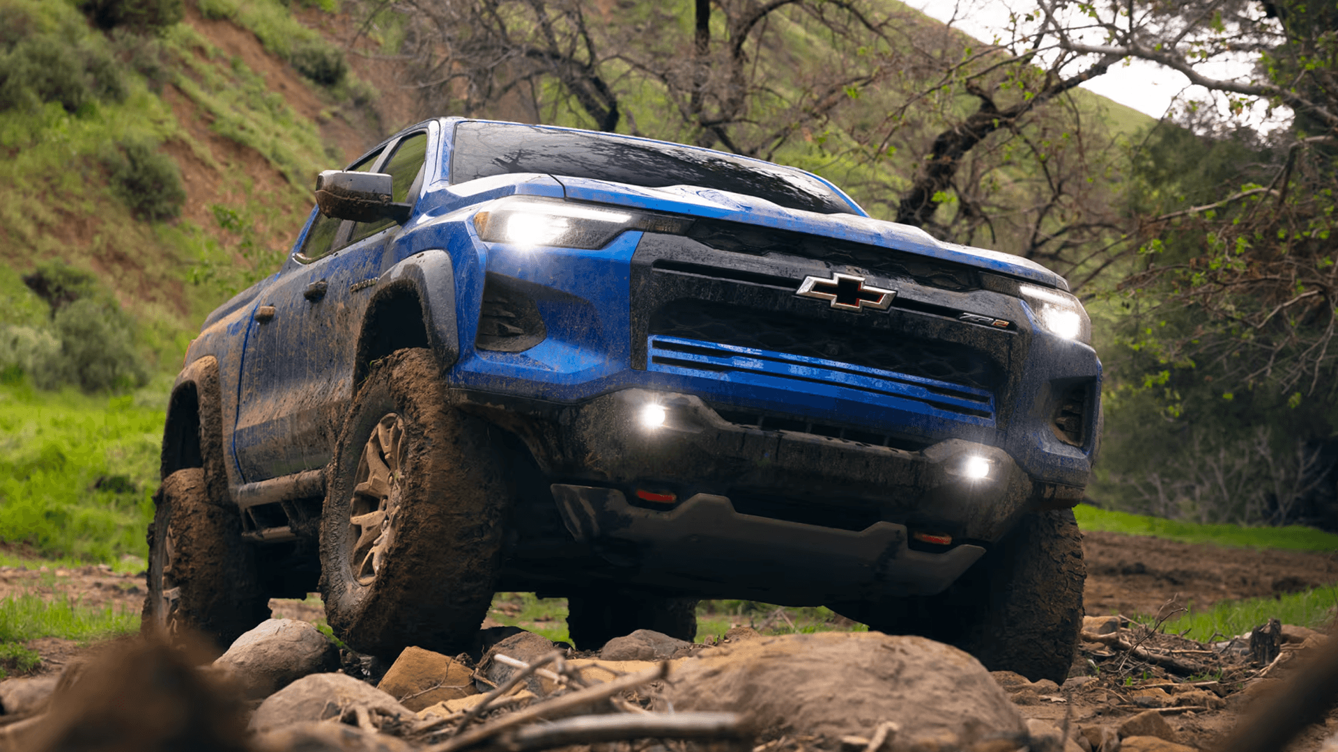Blue Chevy Colorado driving off road over rocks and muddy terrain with bright lighting