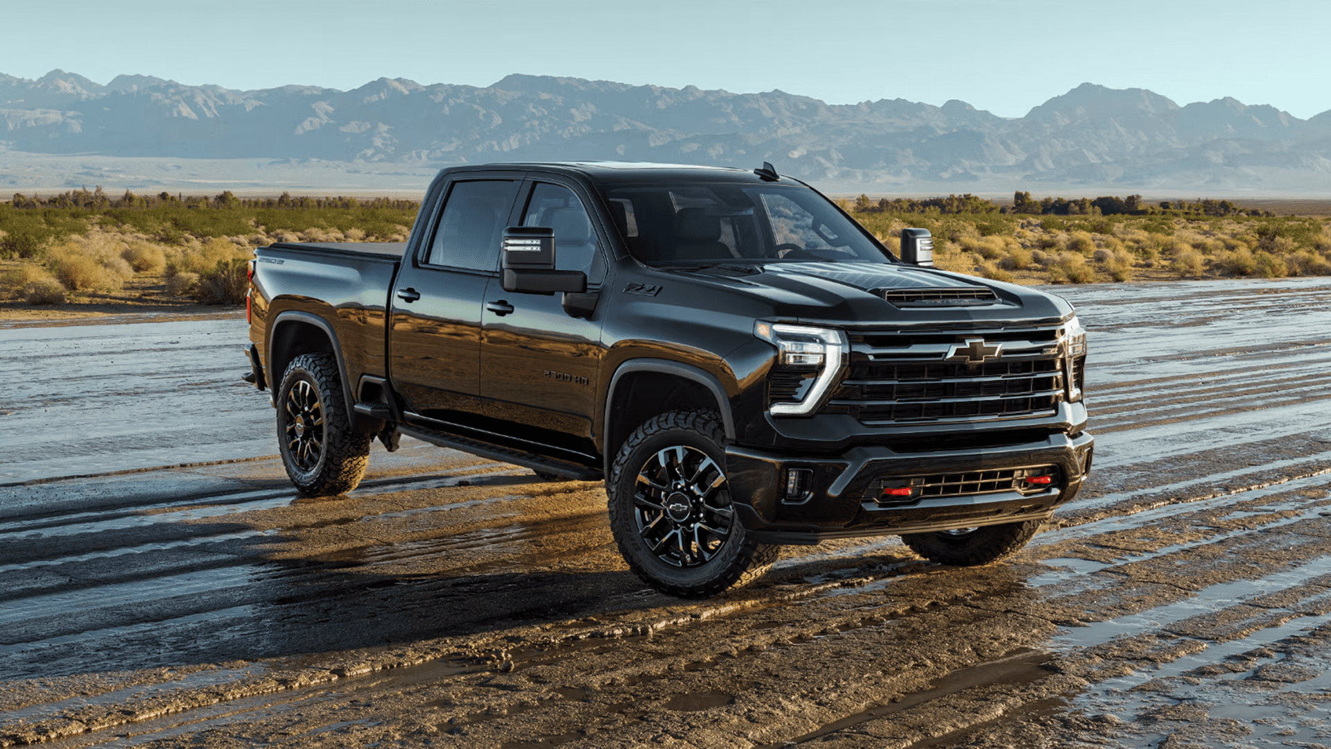 Black 2026 Chevy Silverado HD crew cab parked on desert flats with mountains, showing bold grille, hood scoop, and heavy duty stance