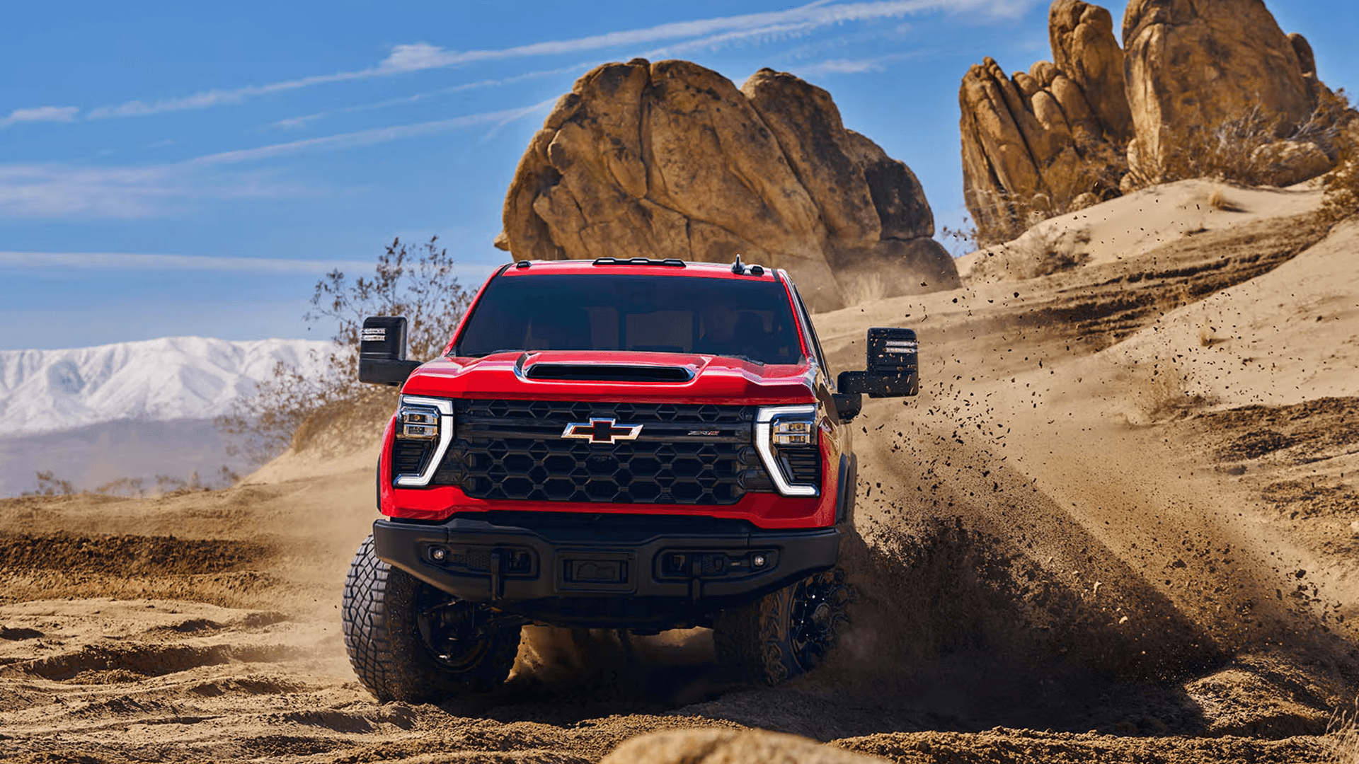 Red 2026 Chevy Silverado HD ZR2 driving through sand, kicking up dirt with off road grille, skid plate area, and wide stance