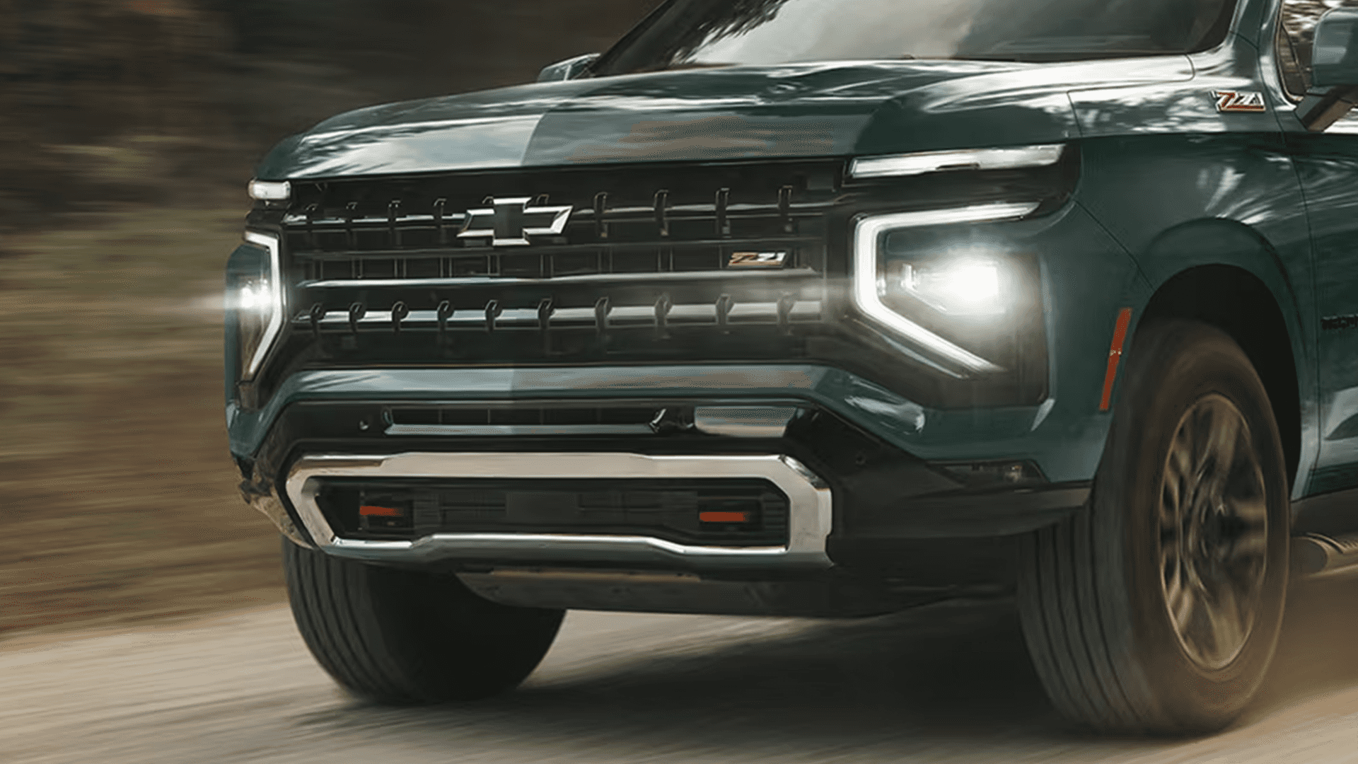 front of a green 2026 Chevrolet Tahoe Z71 showing illuminated headlights and grille driving on a road