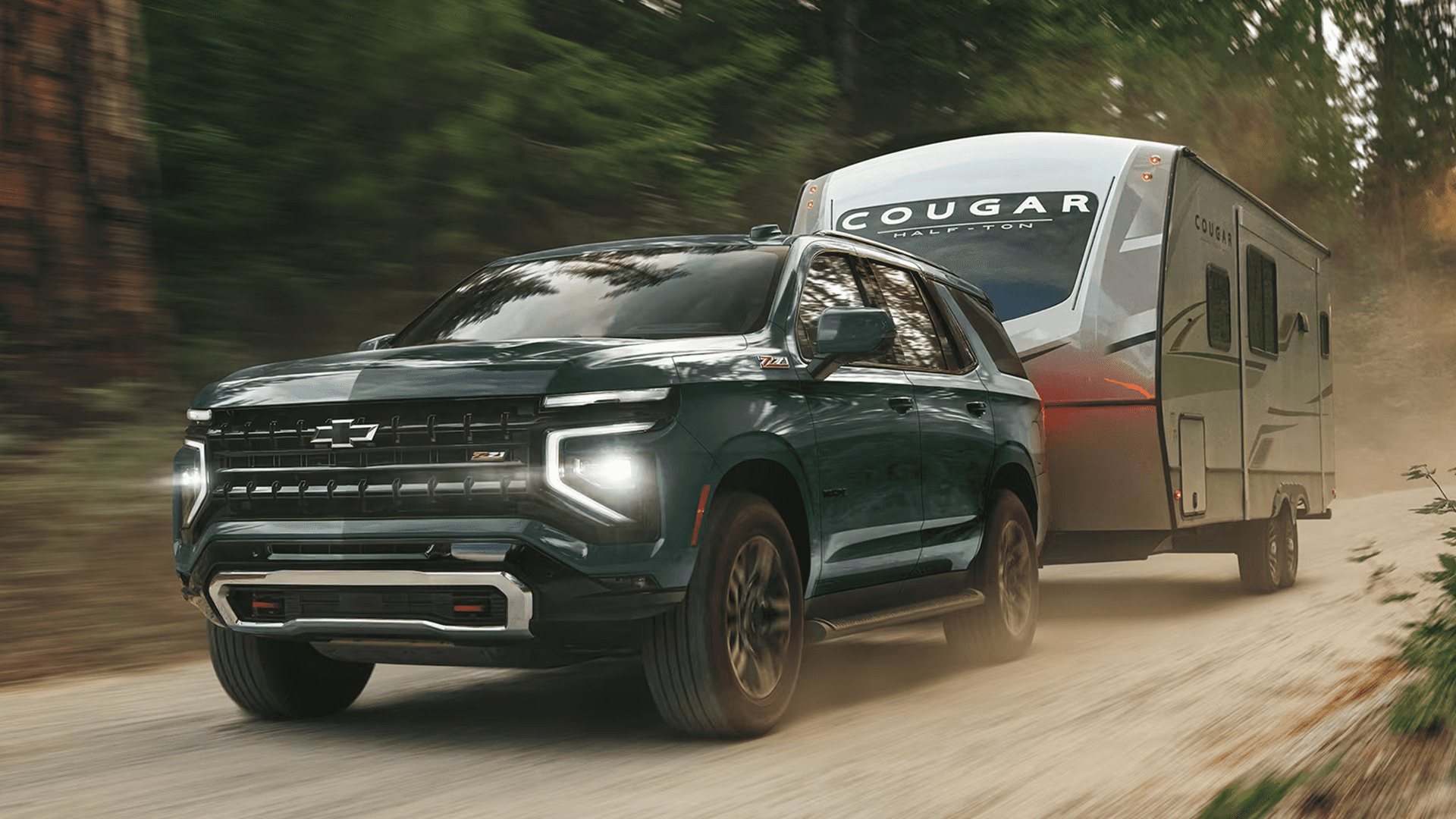 green 2026 Chevrolet Tahoe driving on a dirt road while towing a Cougar travel trailer