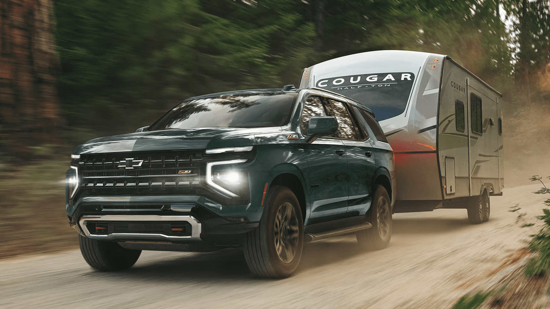2026 Chevy Tahoe towing a Cougar travel trailer on a dirt road through a wooded area