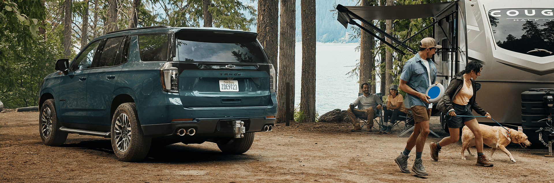 2026 Chevy Tahoe parked at a forest campsite towing a travel trailer while people relax and unload camping gear
