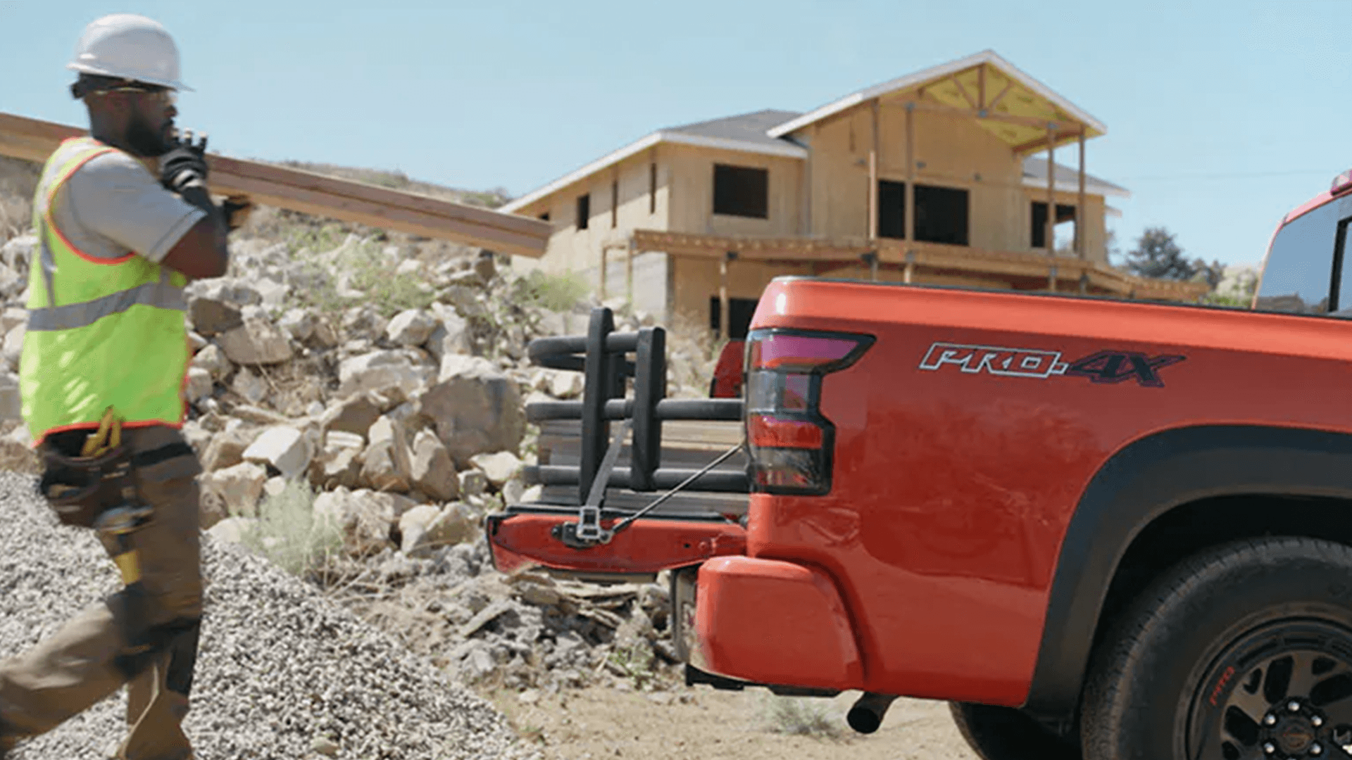 Construction worker carrying lumber beside an orange Nissan Frontier PRO-4X with the tailgate down and a bed extender in place, parked at a home build site