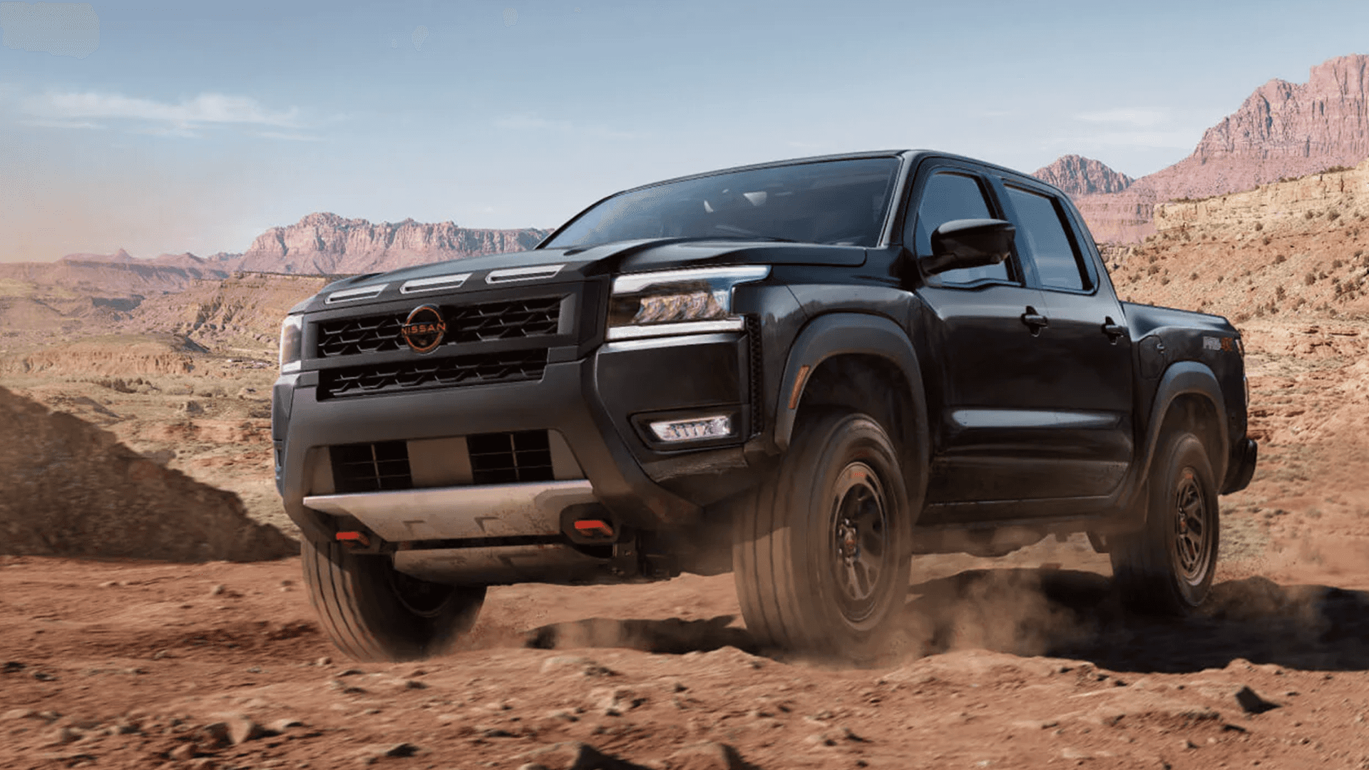 Black Nissan Frontier PRO-4X driving on a desert dirt road with canyon mountains in the background, front three-quarter view showing the bold grille, skid plate, and red tow hooks