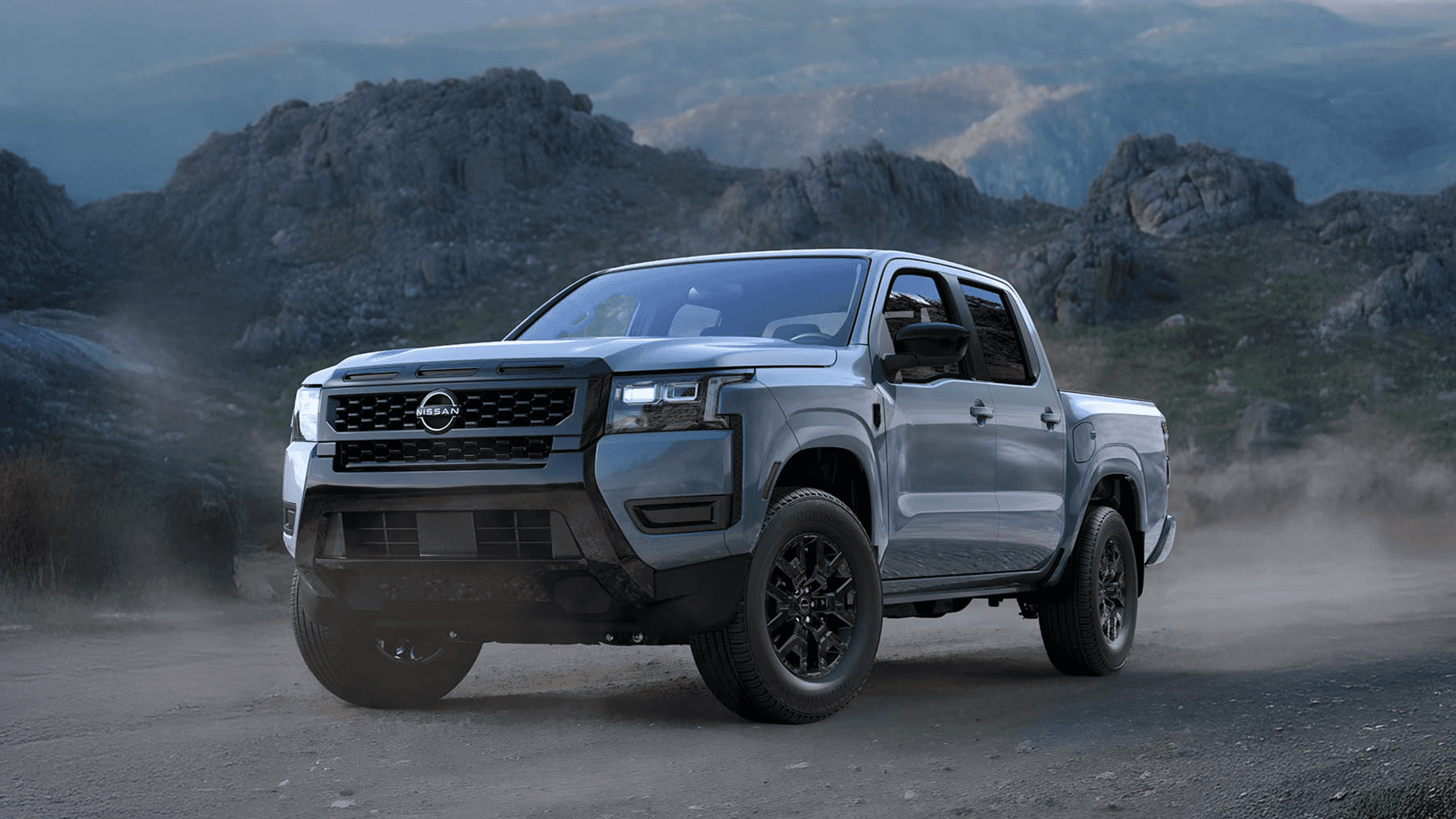 Silver Nissan Frontier driving on a dusty gravel road with rugged mountains behind it, front three-quarter view highlighting the truck’s stance and ground clearance