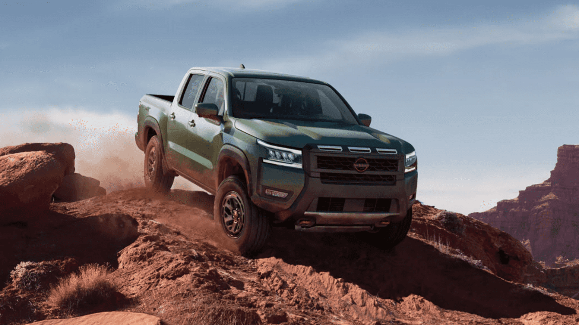 Green Nissan Frontier cresting a red rock ridge, front three-quarter view with dust trailing behind the truck under a wide open sky