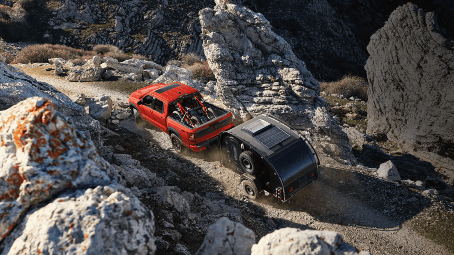 Overhead view of an orange Nissan Frontier towing a compact black camper trailer along a winding rocky mountain road, with gear visible in the truck bed