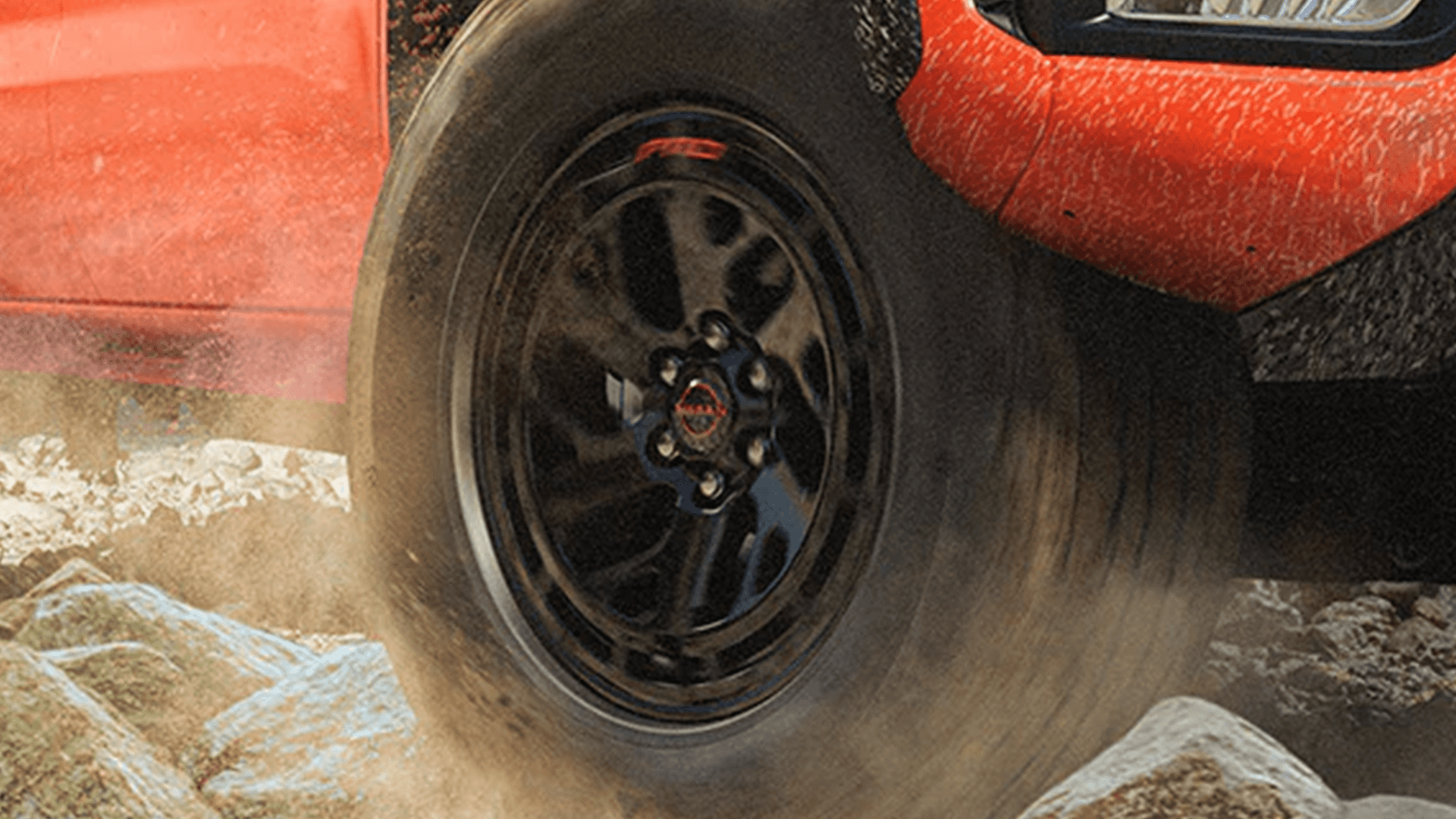 Close-up of a Frontier black alloy wheel and all-terrain tire kicking up dust over rocks, with the red body panel splattered with mud near the wheel arch