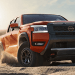 Orange 2026 Nissan Frontier midsize pickup driving on desert terrain with dust trail and mountain backdrop