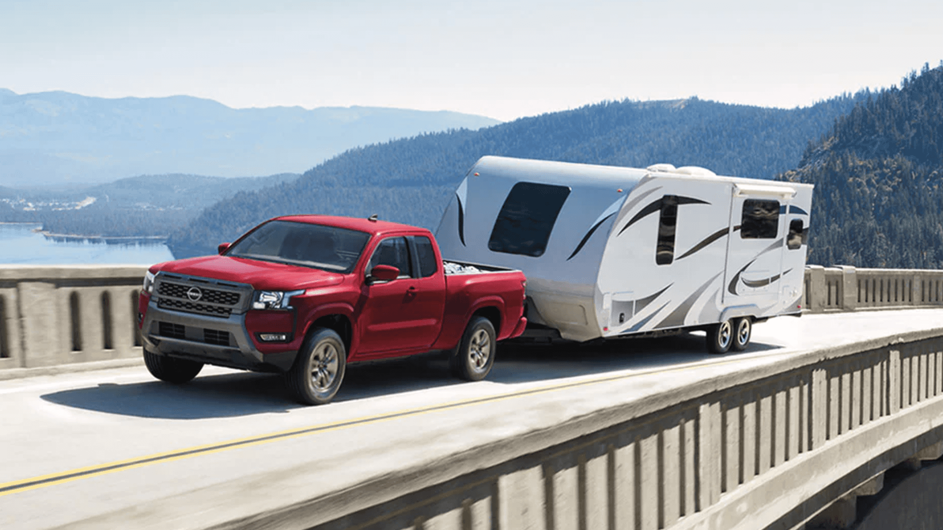 Red Nissan Frontier towing a travel trailer across a bridge with mountains and water in the background