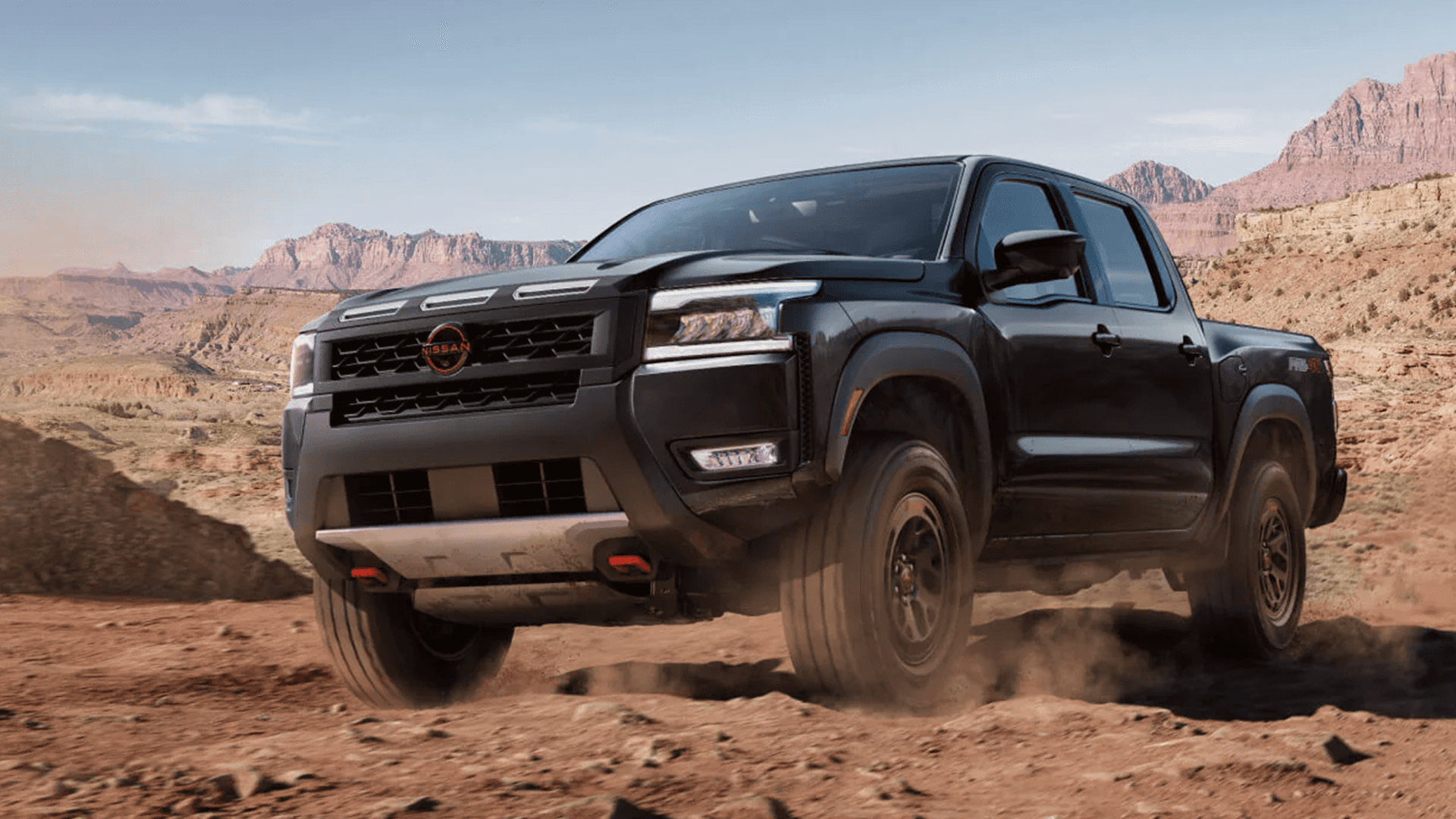Black Nissan Frontier driving on a desert dirt road with red rock cliffs in the background