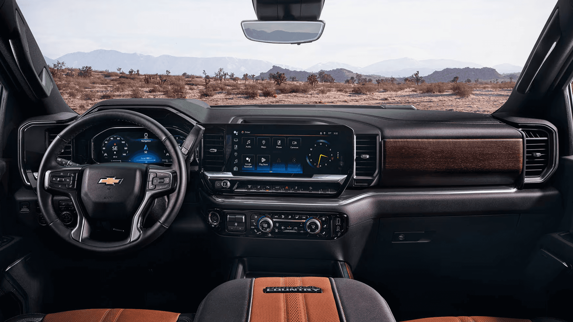 Silverado 2500 HD cabin view showing the steering wheel, digital gauge cluster, wide center touchscreen, wood trim dash, and two tone High Country seats with desert landscape outside