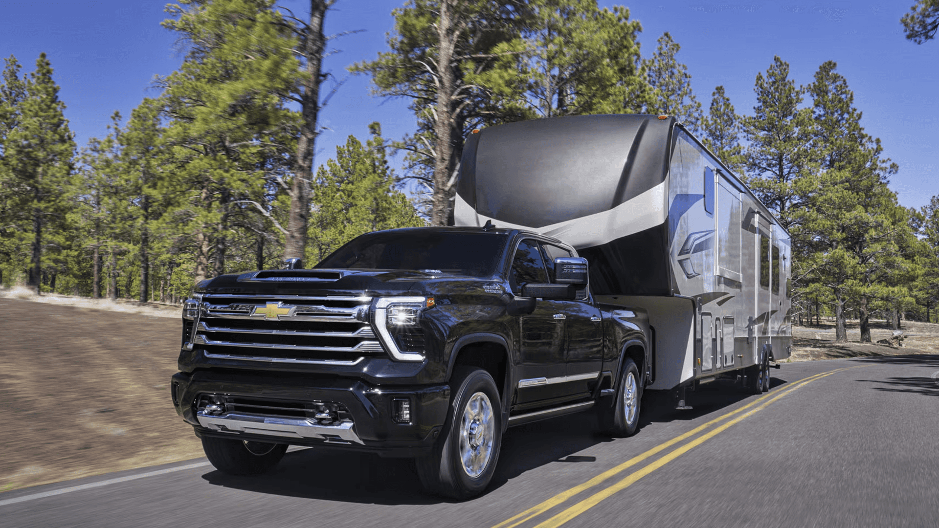 Black Chevy Silverado 2500 HD towing a large fifth wheel camper on a two lane road through pine trees