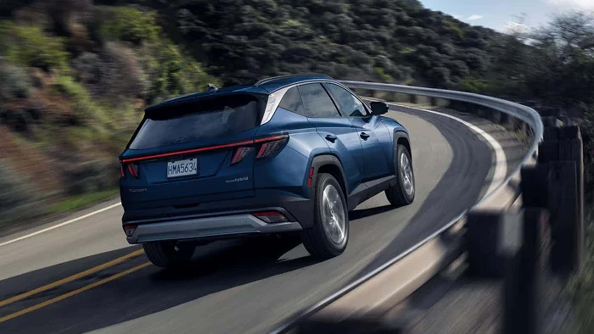 Rear view of a blue Hyundai Tucson Hybrid driving on a winding road, emphasizing stability, handling, and hybrid efficiency.