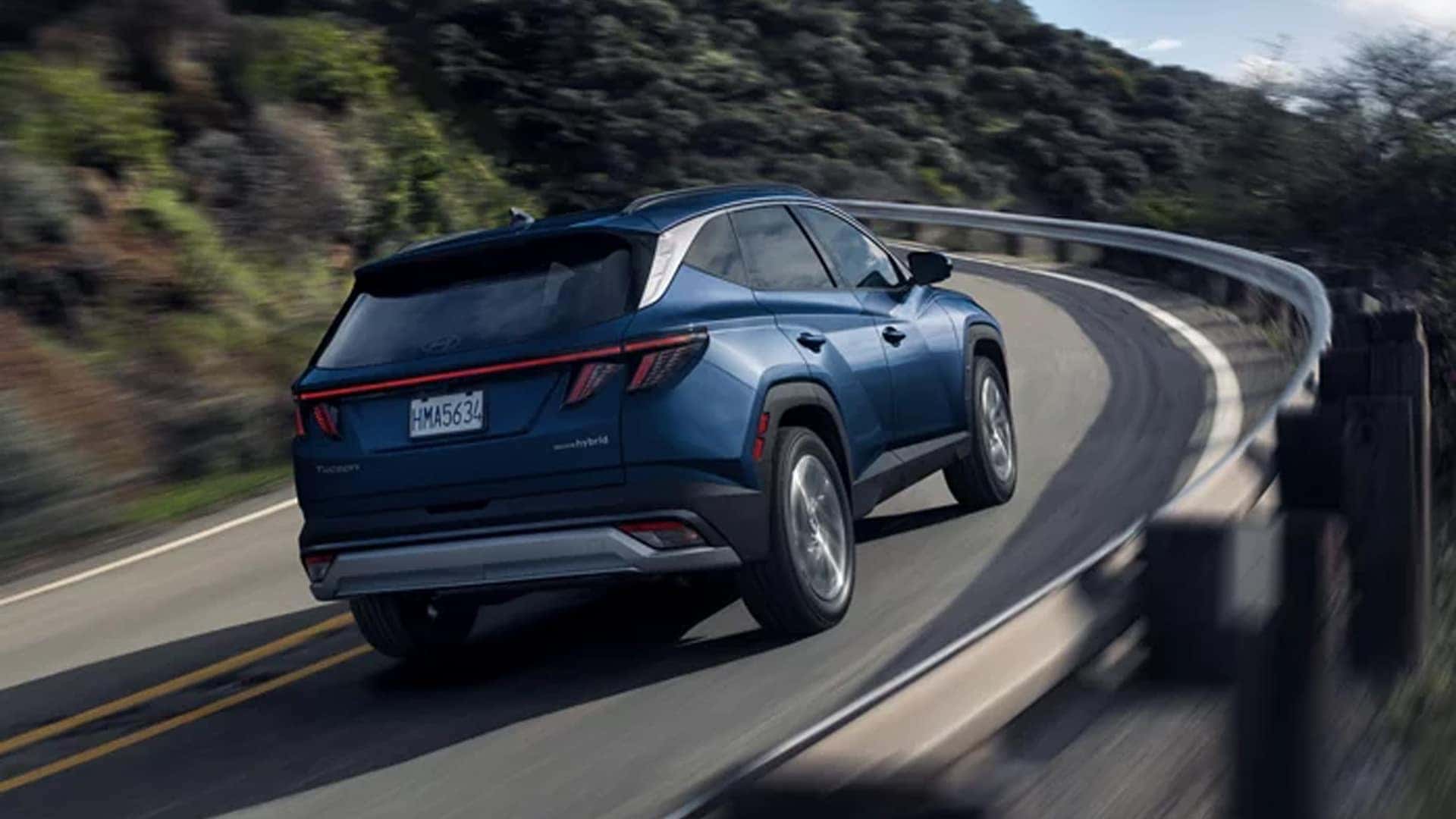 Rear view of a blue Hyundai Tucson Hybrid driving along a winding mountain road, illustrating handling and available HTRAC all-wheel drive capability.