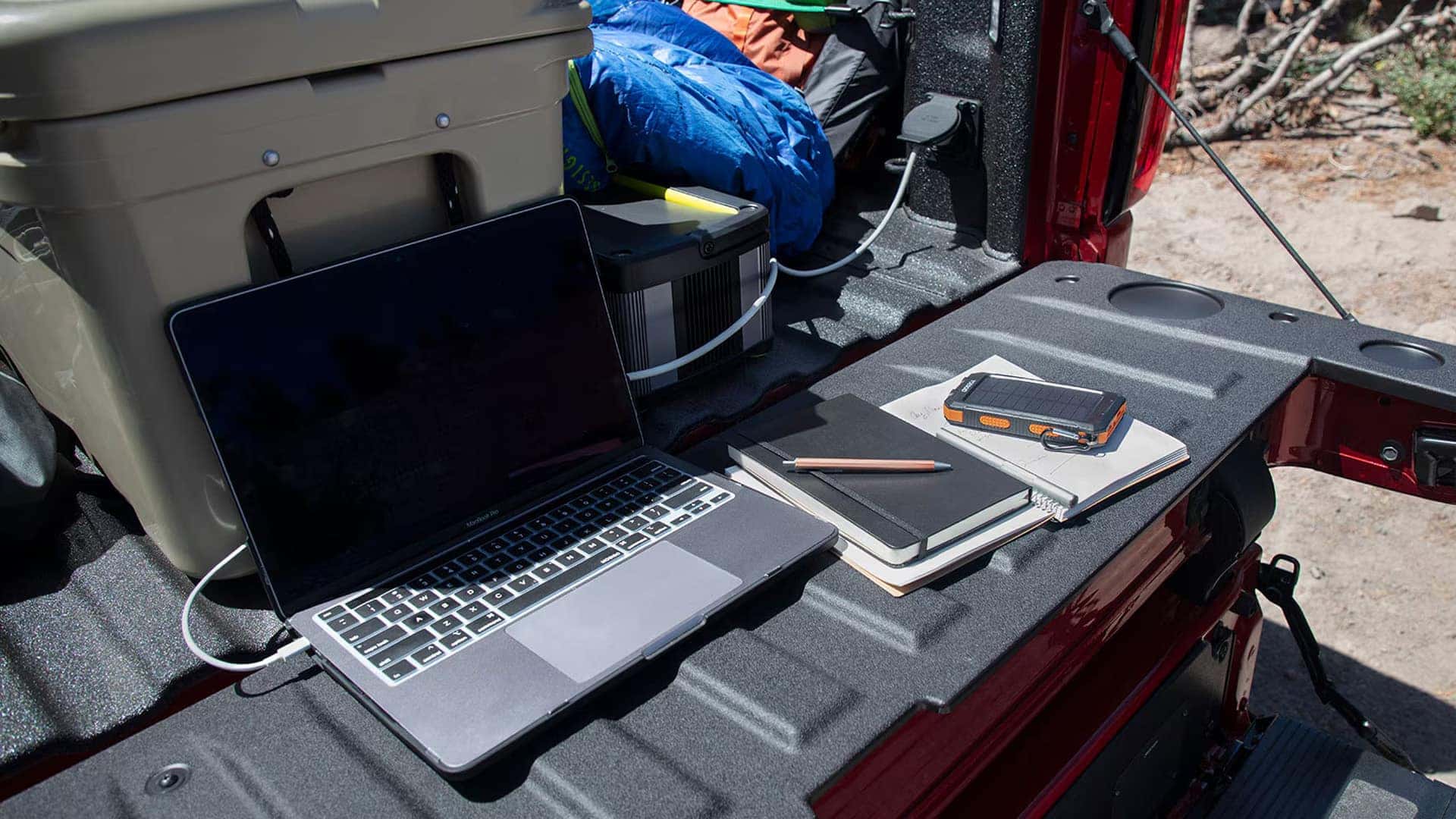 Open Sierra 1500 tailgate used as a work surface with a laptop, notebooks, and gear in the truck bed at an outdoor campsite.