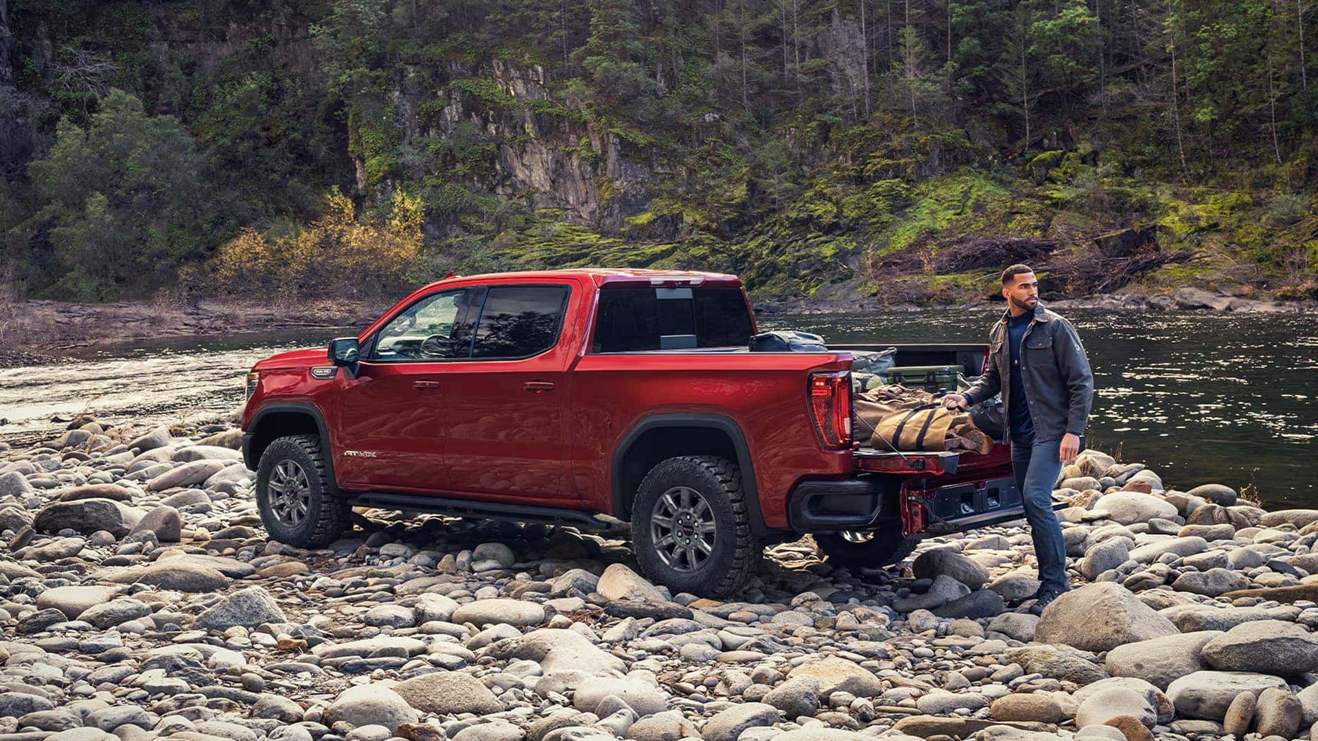 Red 2026 GMC Sierra 1500 AT4X parked near a river with gear loaded, showcasing off-road performance and cargo versatility.