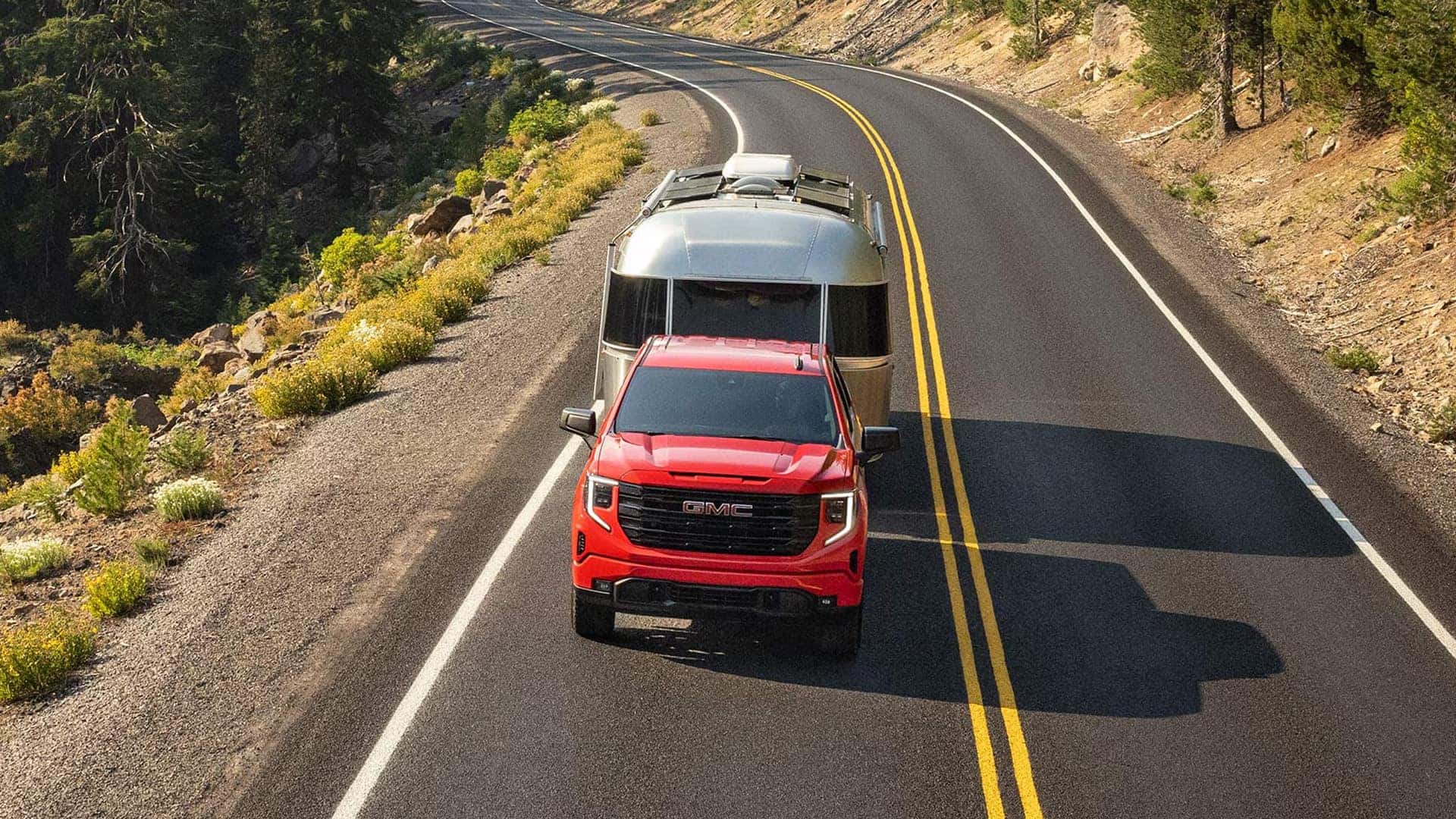 Overhead view of a red 2026 GMC Sierra 1500 towing a travel trailer on a winding two-lane road through rugged terrain.