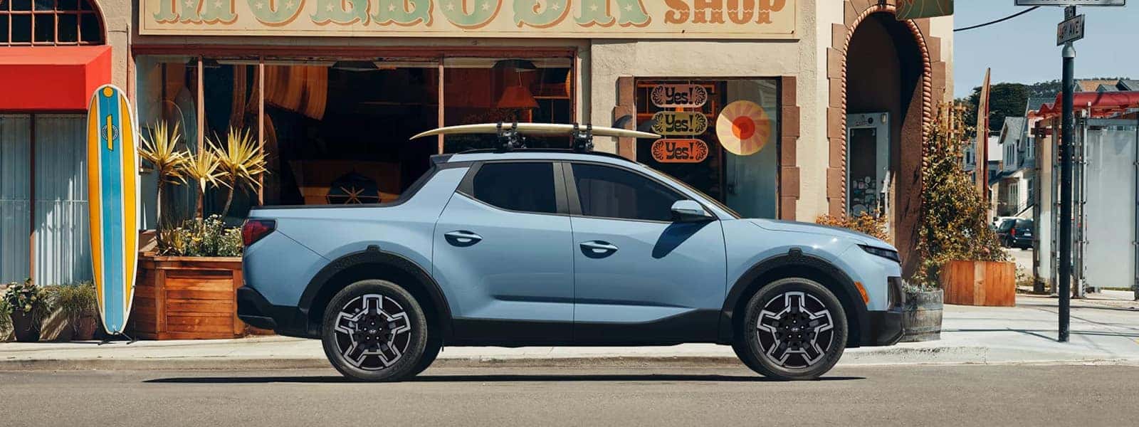 Side profile of a blue 2026 Hyundai Santa Cruz parked on a city street with roof rack and surfboard mounted on top.