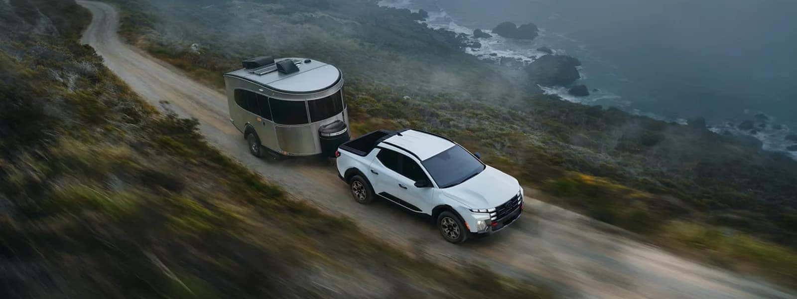 2026 Hyundai Santa Cruz towing a compact travel trailer along a coastal road with ocean cliffs in the background