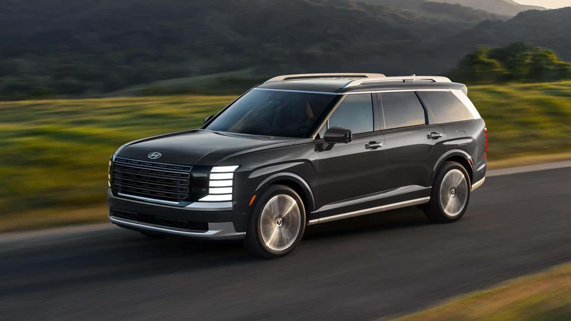 Front three quarter view of a 2026 Hyundai Palisade Hybrid driving on an open road, highlighting its large three row SUV design and hybrid powertrain.