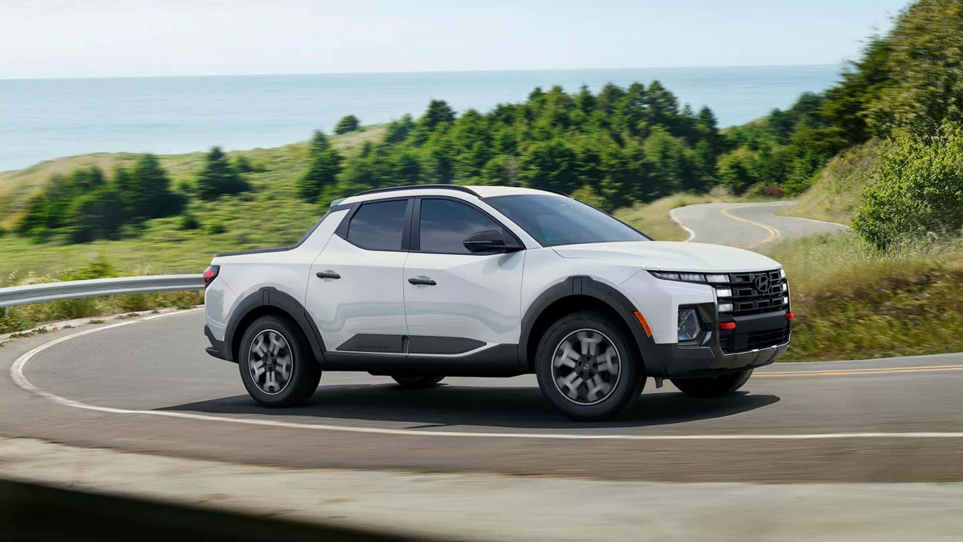 White 2026 Hyundai Santa Cruz driving on a winding coastal road, showing compact pickup shape and bold front grille design.