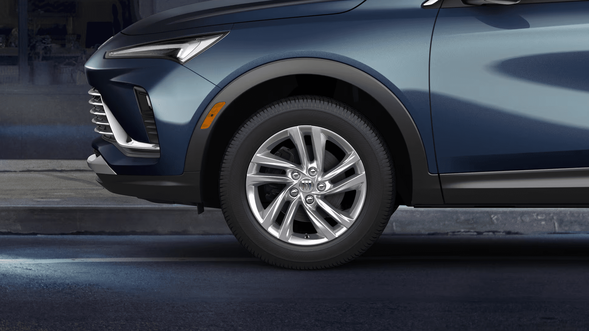 Close up of the front driver side wheel area on a blue 2026 Buick Envista, showing a bright silver multi spoke alloy wheel, tire tread, black wheel arch cladding, and the lower front fascia
