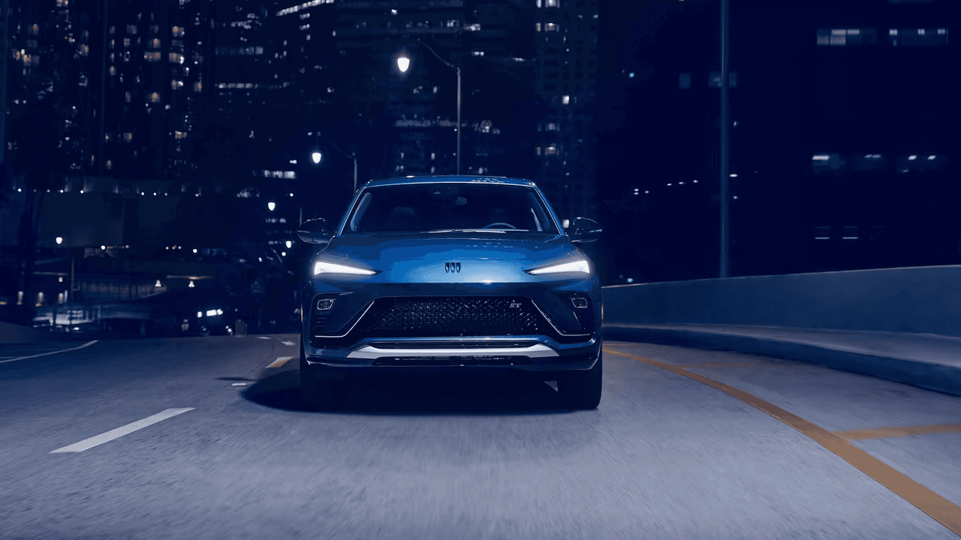 Front view of a blue 2026 Buick Envista driving at night on a city roadway, with LED headlamps illuminated and the wide grille centered beneath streetlights and high rise buildings