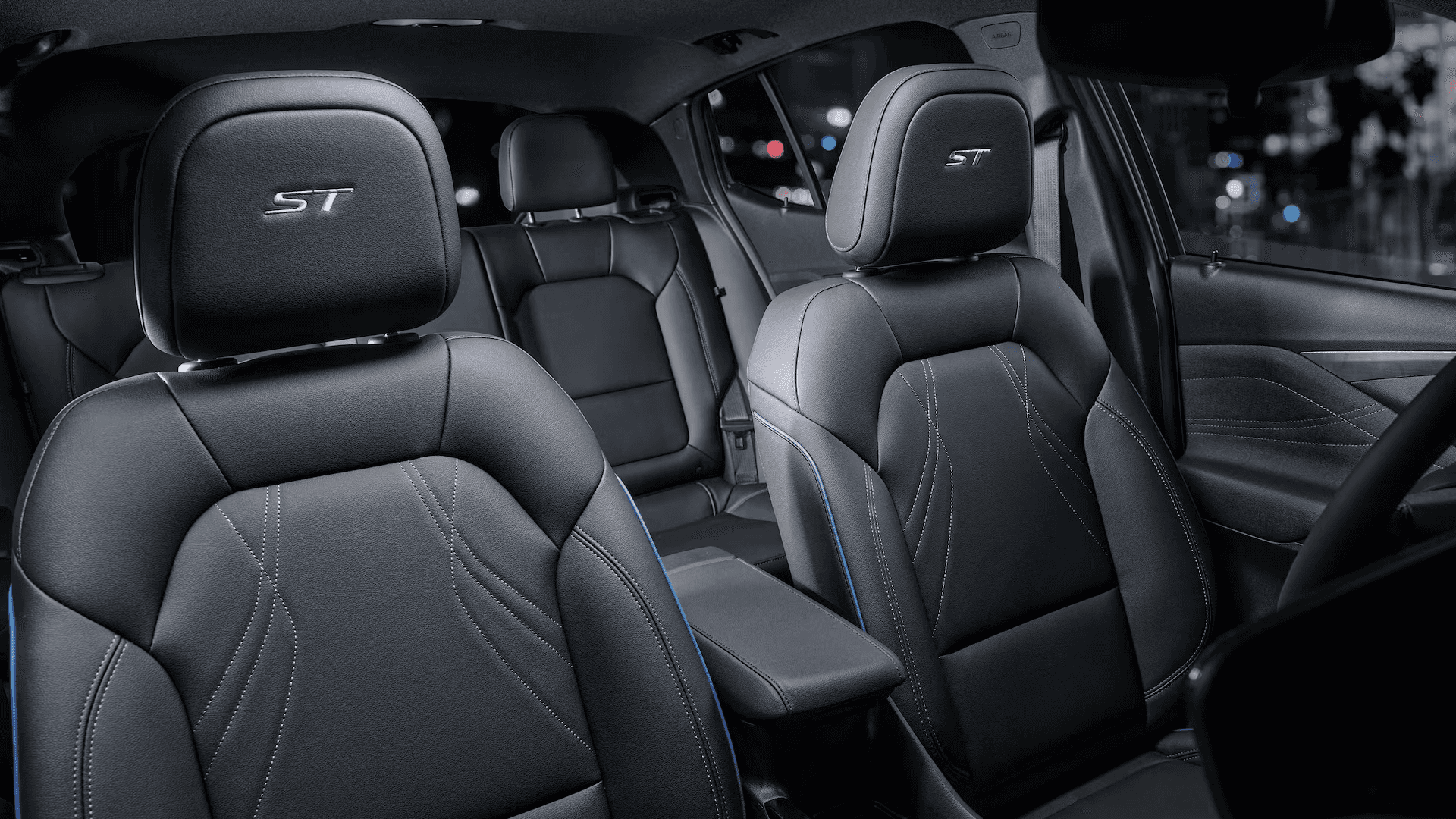 Front cabin view of a Buick Envista with black seats featuring ST embroidery on the headrests, visible contrast stitching, center armrest, and second row seating in the background with city lights outside the windows.