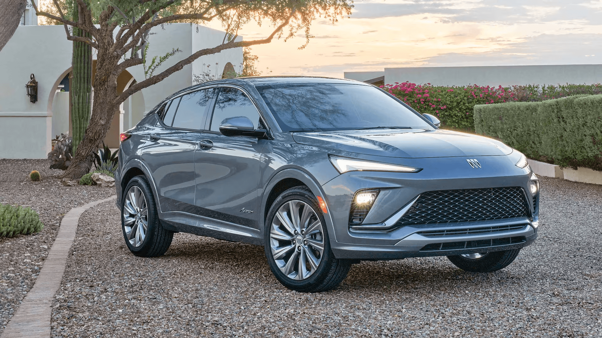 Gray Buick Envista parked on a gravel driveway at sunset near a desert style home with a cactus and flowering landscaping, shown from a front three quarter angle with the grille and LED lighting visible.