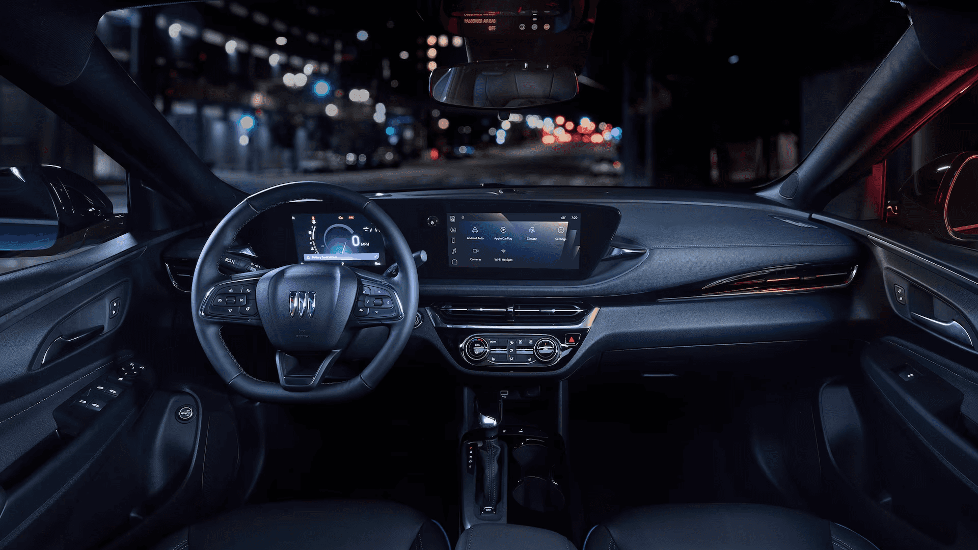Driver’s view of the Buick Envista cockpit at night showing the steering wheel, digital instrument cluster, center touchscreen display, climate controls, and dashboard trim with blurred city streetlights beyond the windshield.