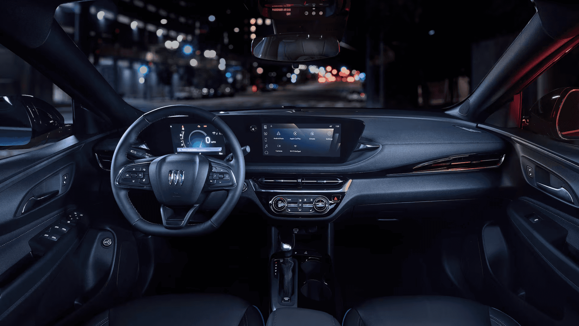 Driver-facing view of the 2026 Buick Envista cockpit at night showing the steering wheel, digital instrument cluster, wide infotainment display, climate controls, and illuminated city street lights through the windshield