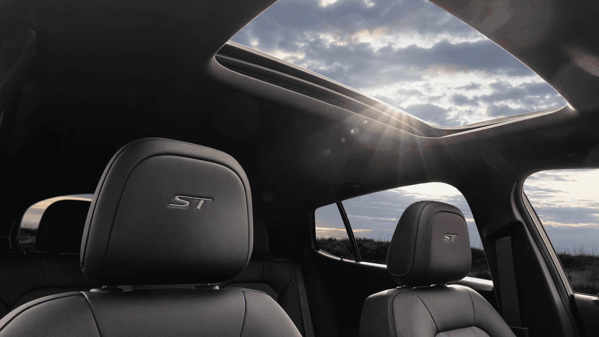 Interior view of the 2026 Buick Envista showing an open power moonroof with sunlight streaming in, with front seat headrests featuring ST logos beneath a dark headliner