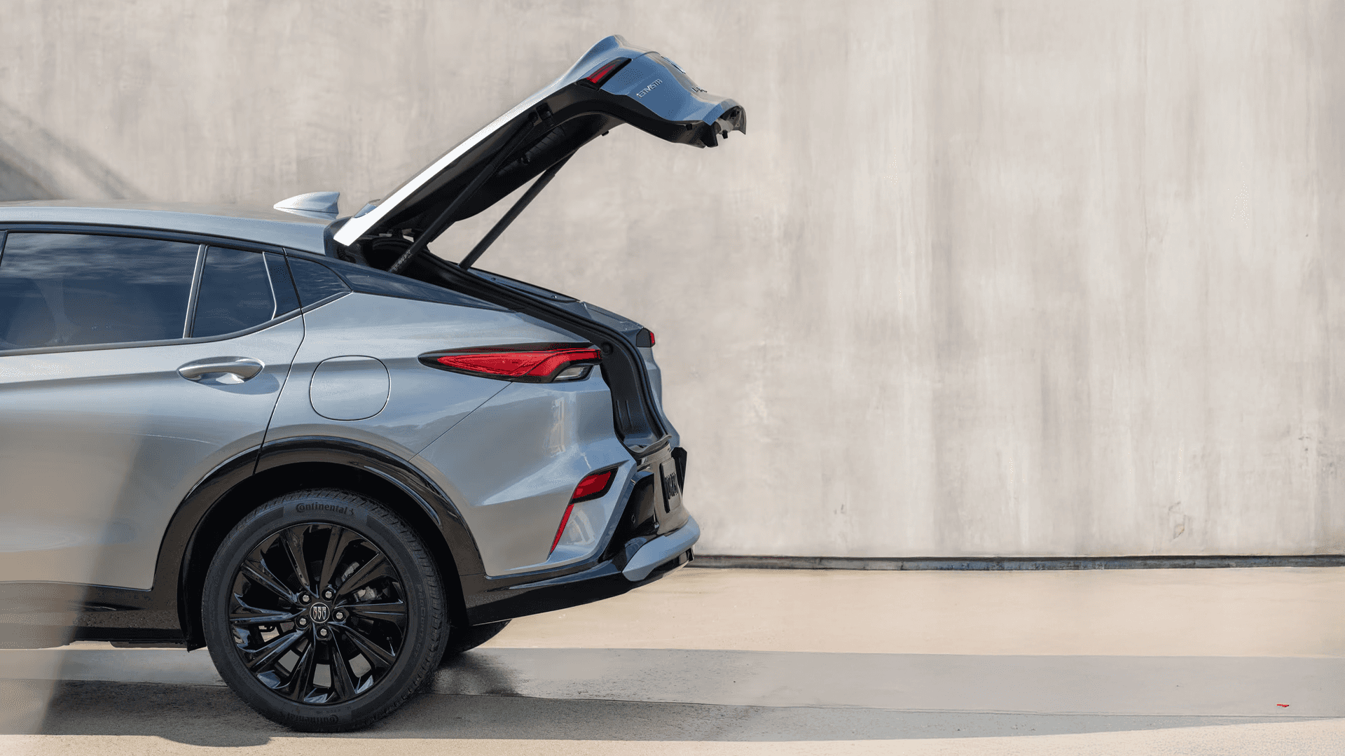 Side rear exterior view of the 2026 Buick Envista with the power liftgate raised, highlighting the rear quarter panel, taillamp, black wheel, and cargo opening area against a concrete wall backdrop