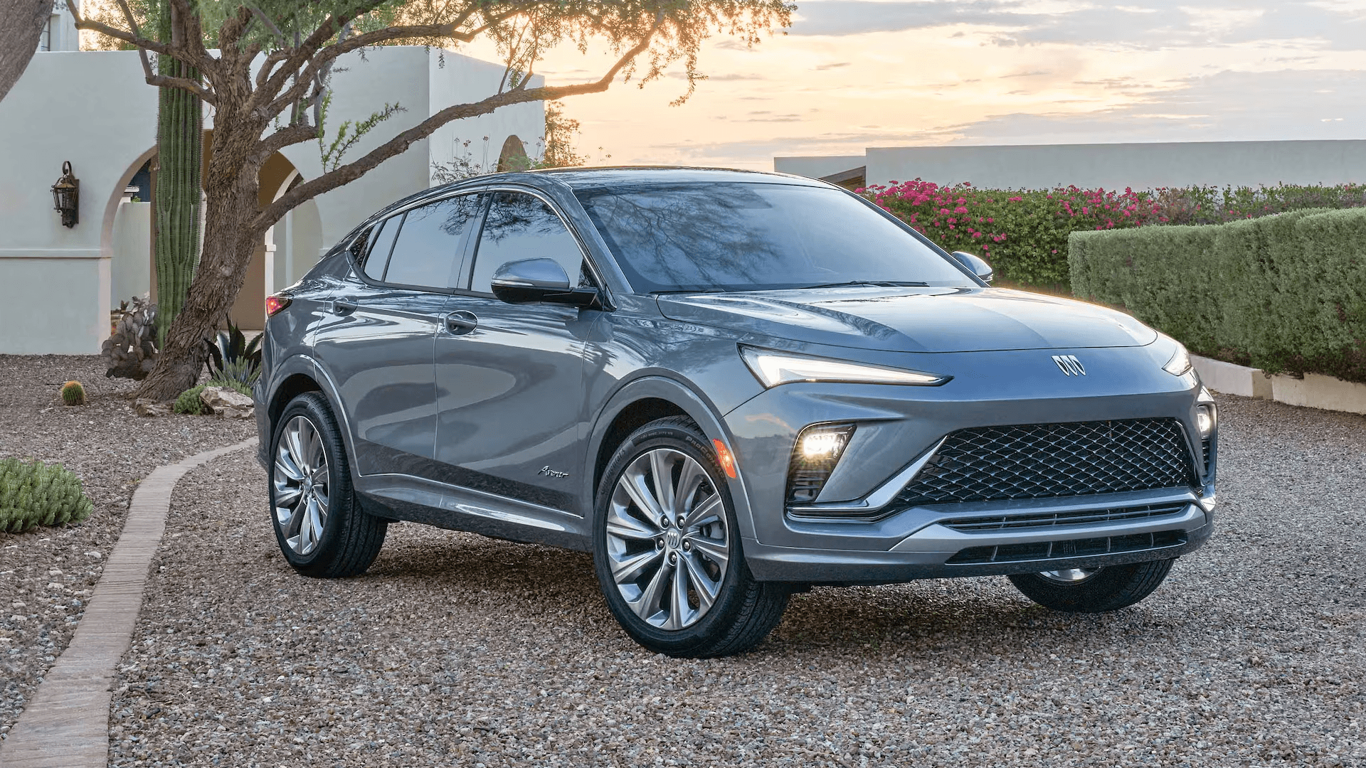 Gray 2026 Buick Envista shown in a front three-quarter view parked on a gravel driveway, with a modern home, desert landscaping, and sunset sky behind it.
