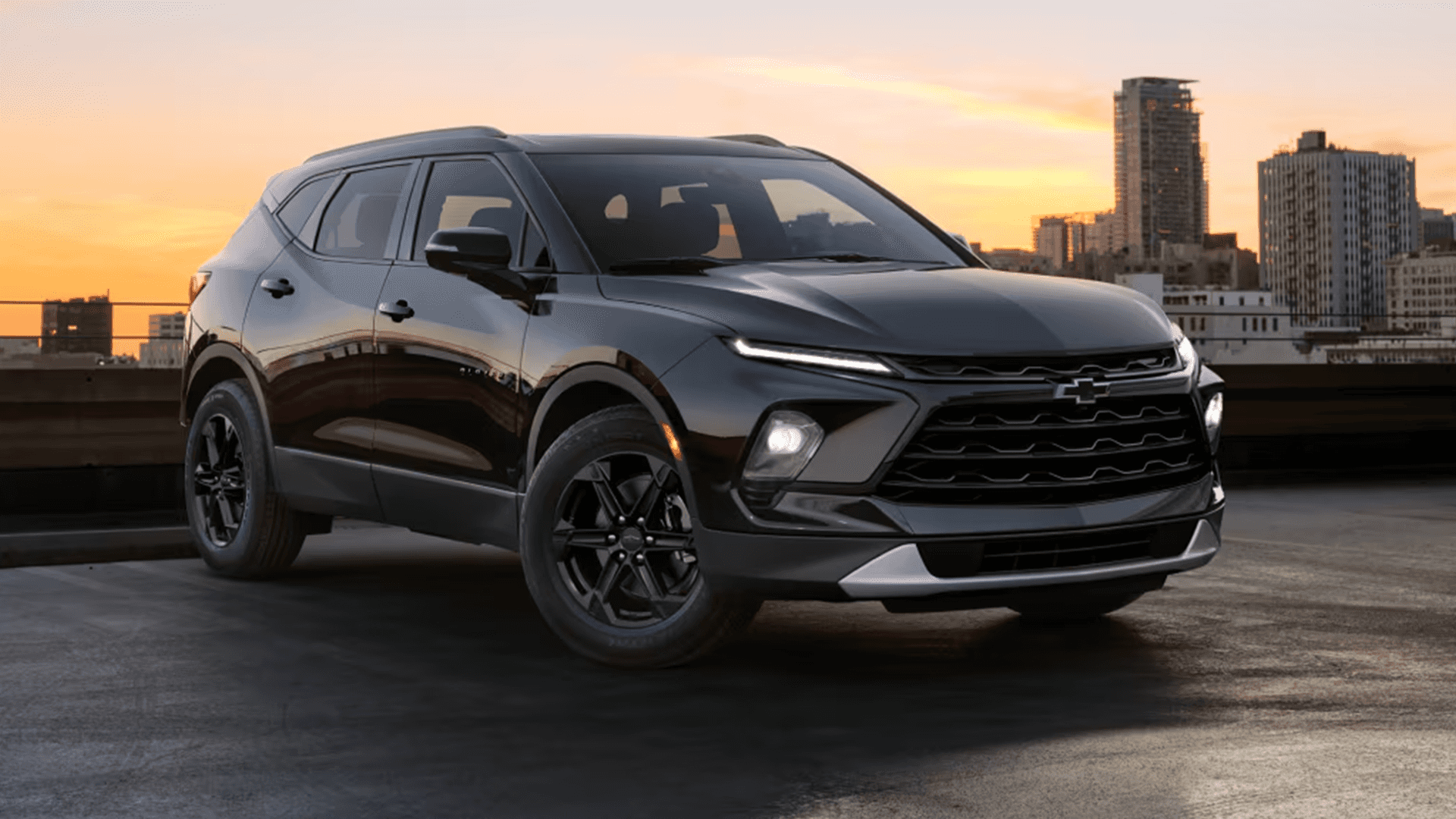 Dark gray 2026 Chevy Blazer parked on a rooftop at sunset, shown from a front three quarter angle with city buildings in the background.