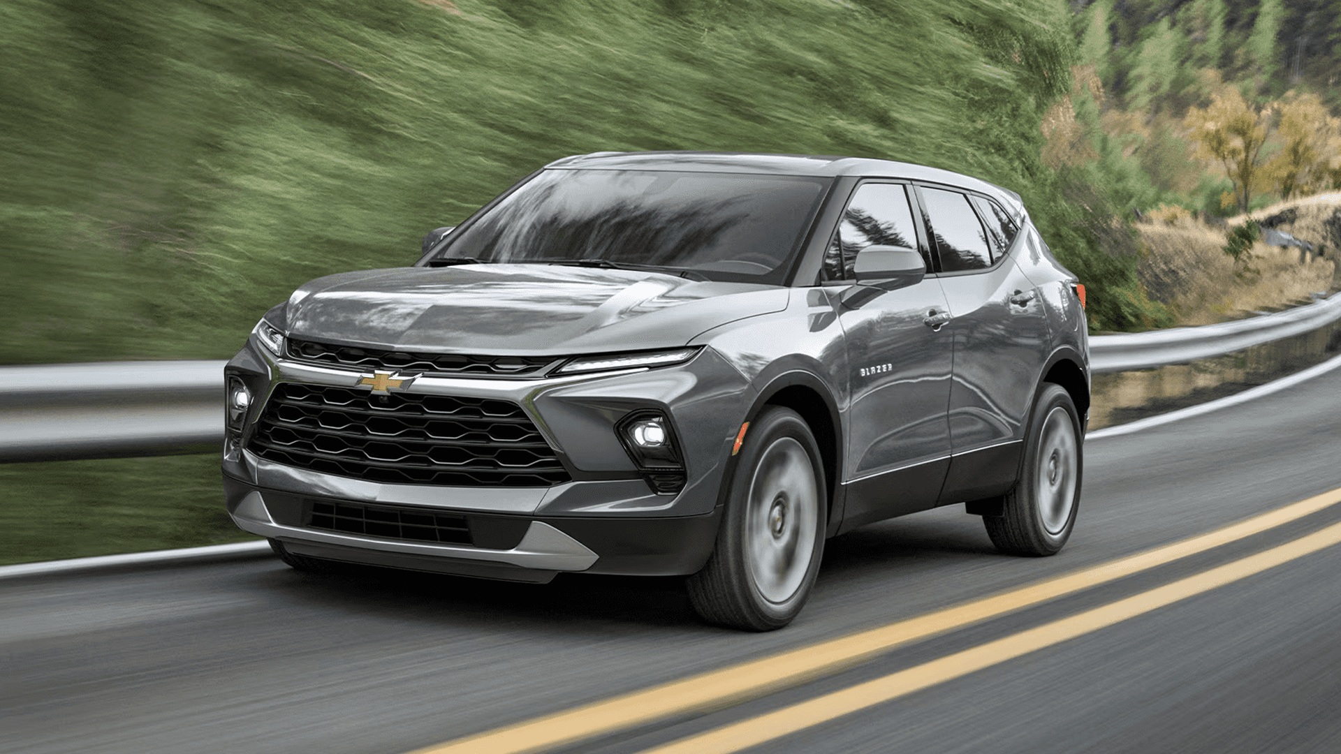 Silver 2026 Chevy Blazer driving on a curved road, shown from a front three quarter angle with surrounding greenery.