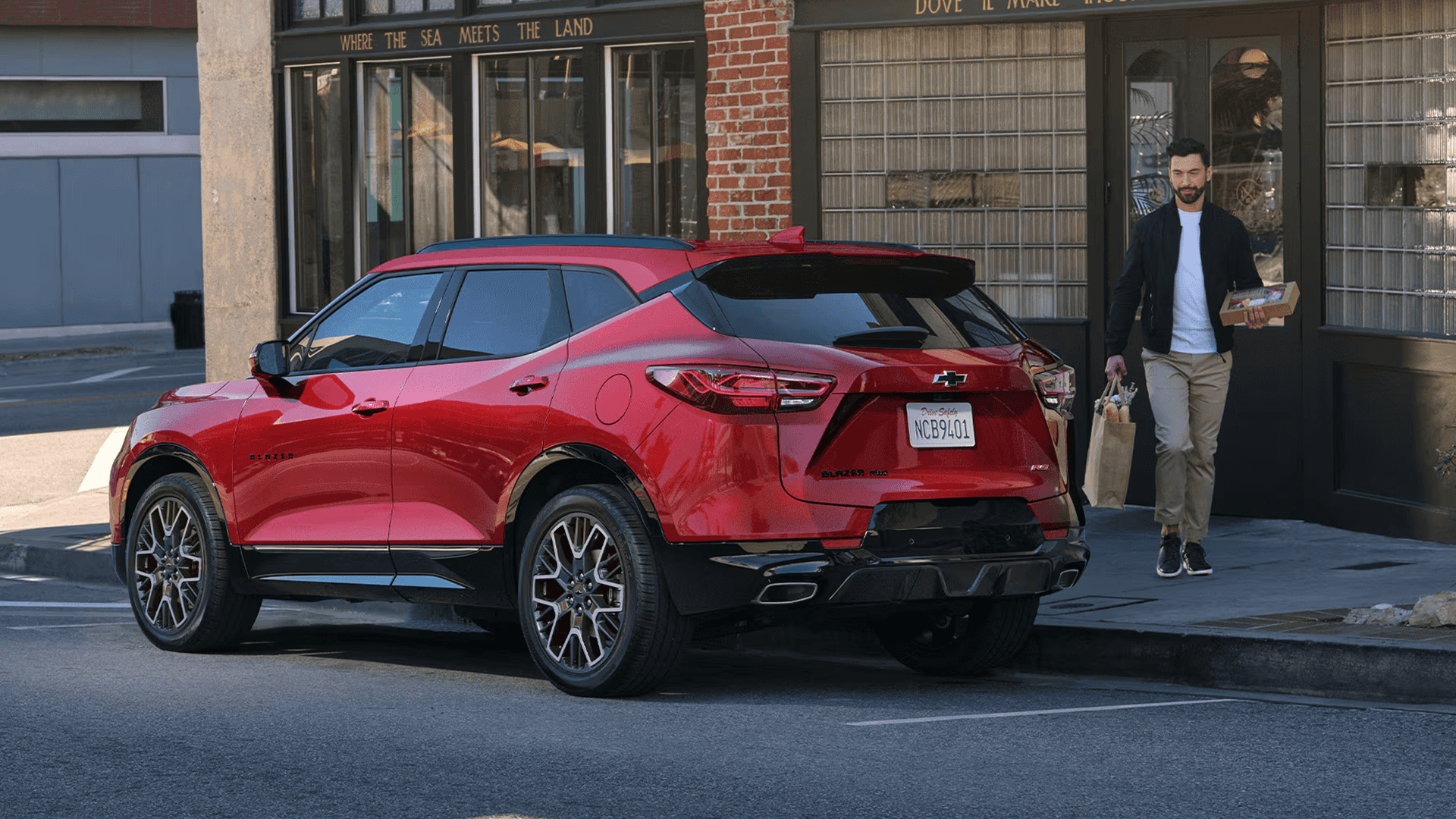 Rear three quarter view of a red 2026 Chevy Blazer parked on a city street, showing rear body lines, taillights, and roof spoiler.