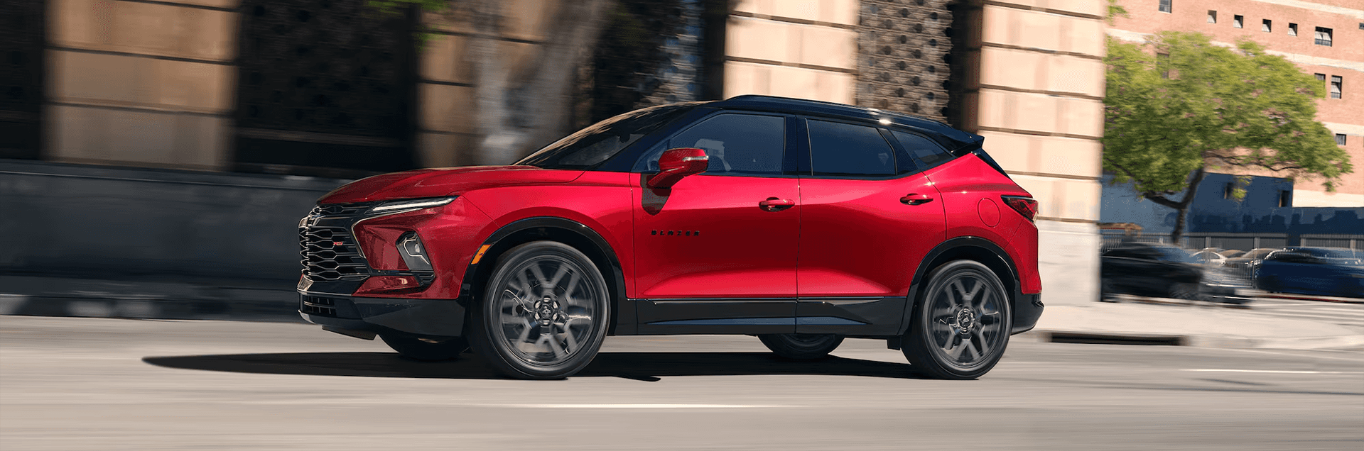Side profile view of a red 2026 Chevrolet Blazer driving in an urban setting, showing the sporty stance, black roof treatment, and wheel design.