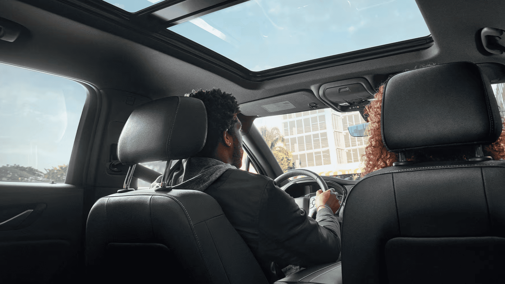 Interior view from the second row of the 2026 Chevy Blazer showing the backs of the front seats, a large panoramic sunroof overhead, and two people seated in the front looking ahead through the windshield.