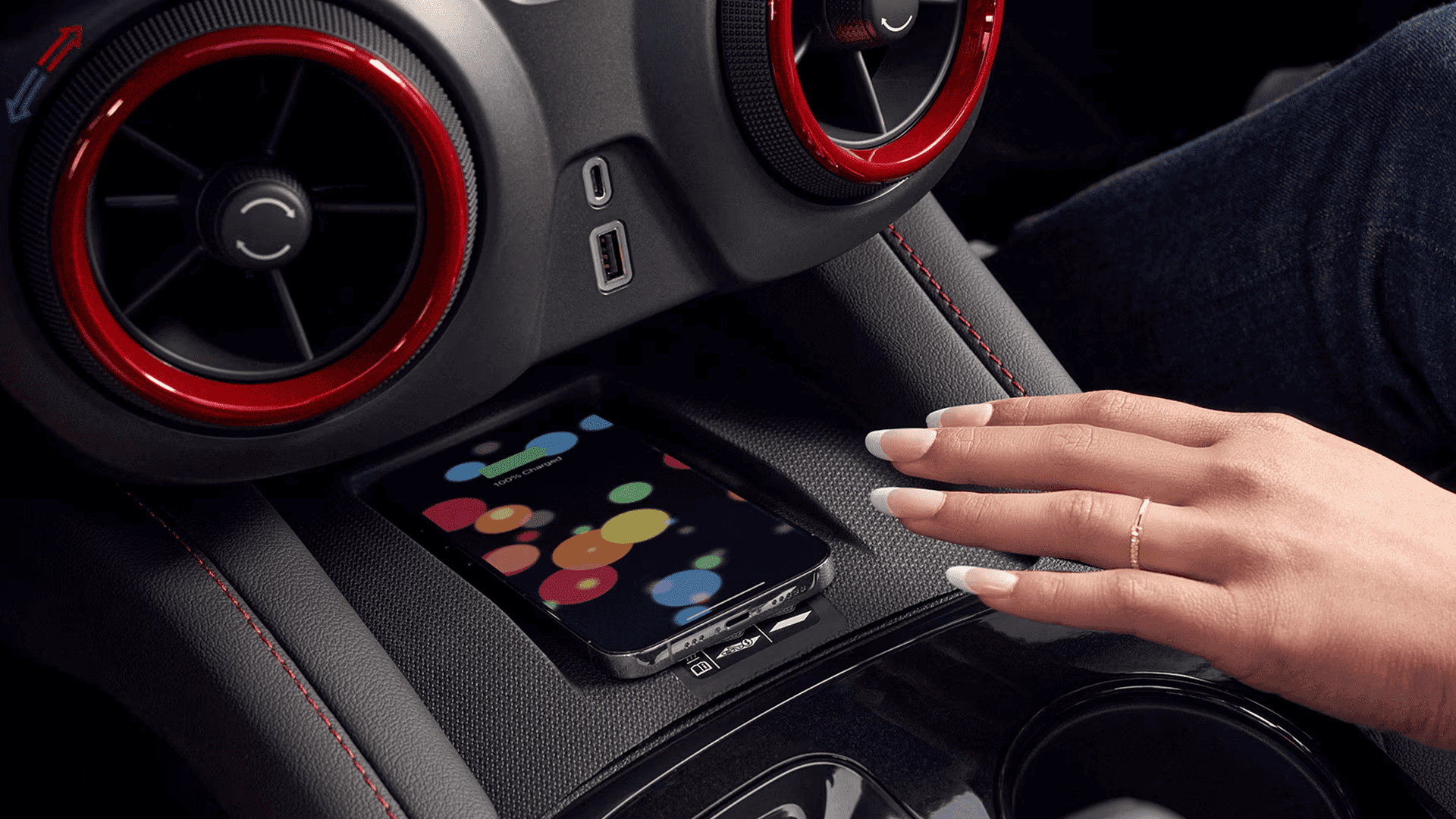 Close-up of a smartphone resting on the wireless charging pad in the 2026 Chevy Blazer, with a hand hovering above it, two red-trimmed circular air vents visible, and USB ports mounted nearby.