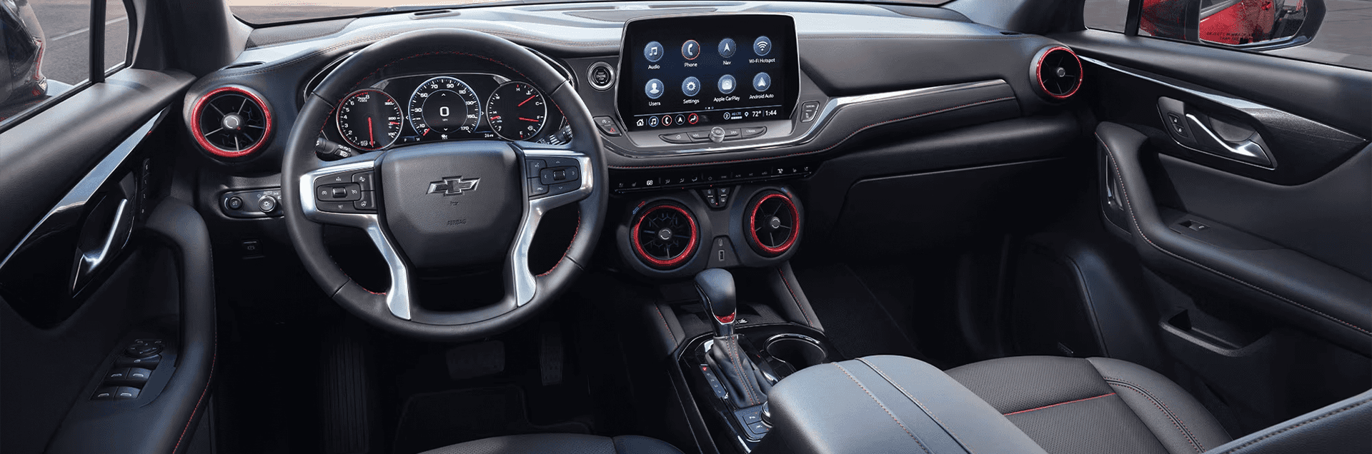 Wide front cabin view of the 2026 Chevy Blazer showing the steering wheel, gauge cluster, center touchscreen, circular air vents with red trim, gear shifter, dual cupholders, and black seats with red stitching.