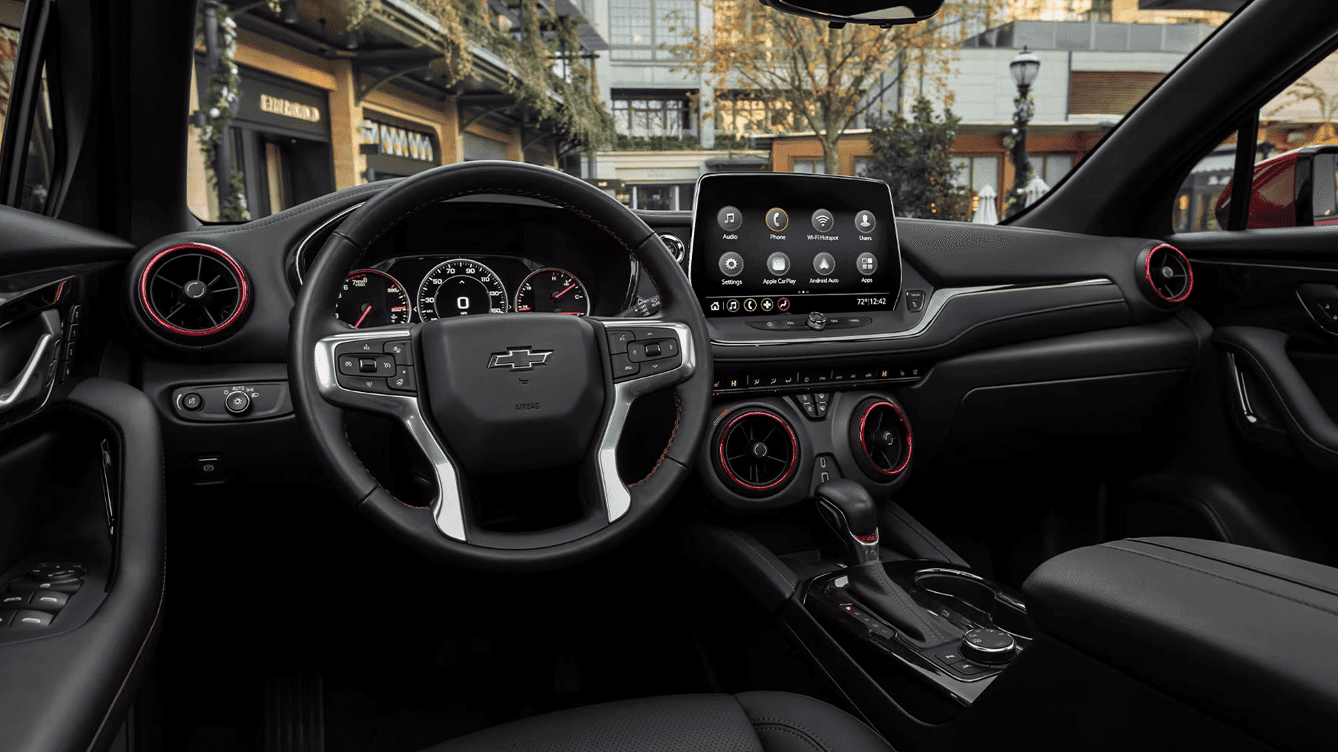 Wide interior view of a Chevy Blazer cockpit showing the steering wheel, analog gauge cluster, and center touchscreen with menu icons including Audio, Phone, Wi Fi Hotspot, Apple CarPlay, and Android Auto, with red accented circular air vents.