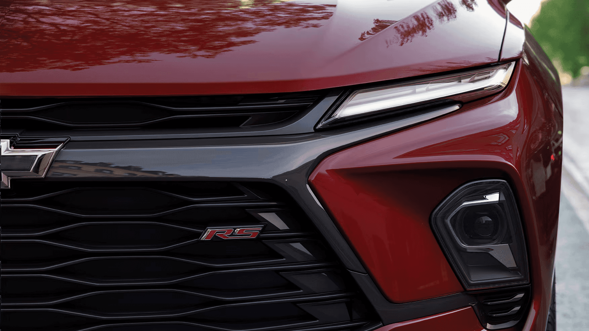 Close view of the front end of a red Chevy Blazer showing the headlamp, grille with RS badge, and front corner camera area, with reflections of trees across the hood.