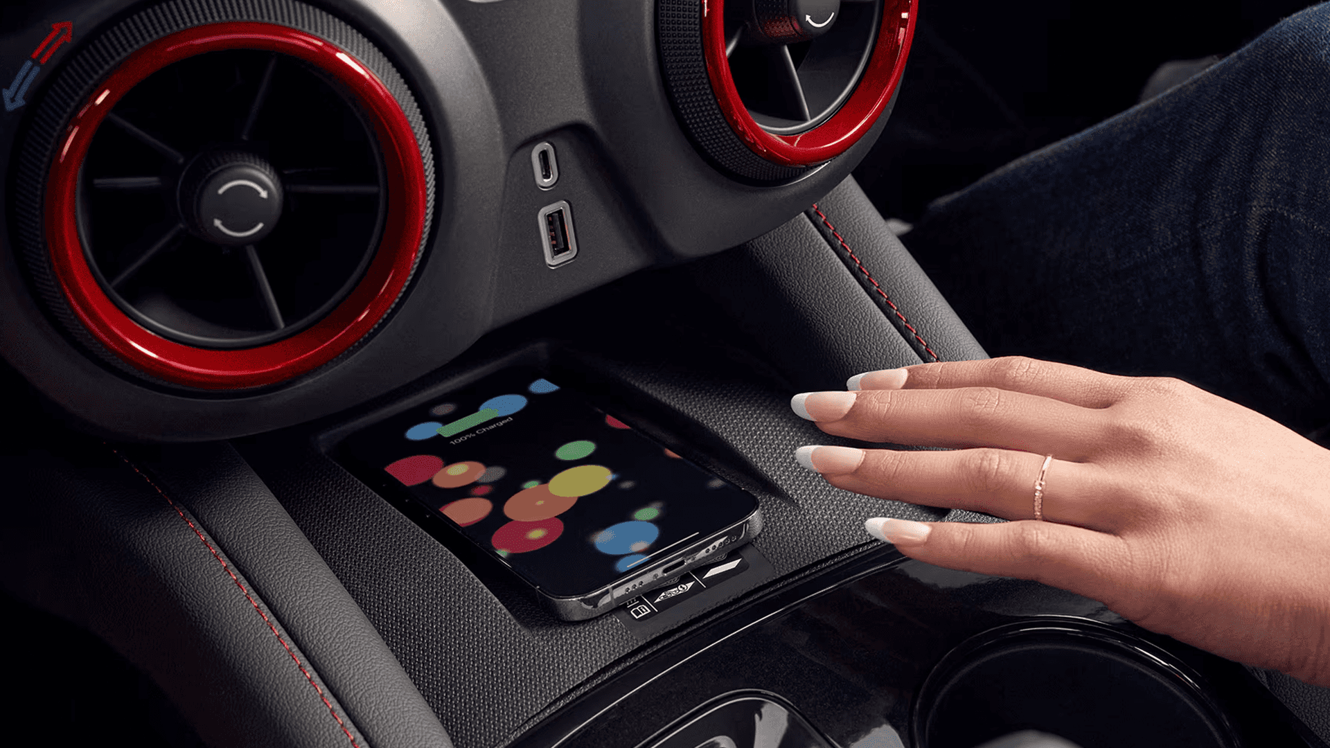Close view of the center console showing a smartphone resting on the wireless charging pad, a hand hovering above it, and nearby USB C and USB A ports beneath red accented circular air vents.