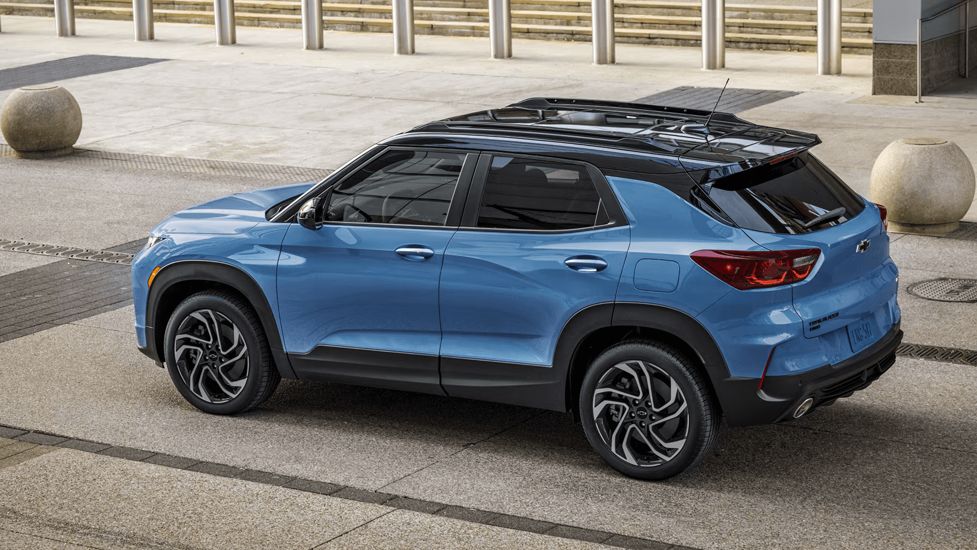 Overhead rear three quarter view of a blue Chevy Trailblazer with a black roof parked on a paved plaza near concrete posts.