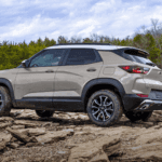 Rear three quarter view of a light tan Chevy Trailblazer positioned on uneven rocky terrain near a lake and wooded shoreline.