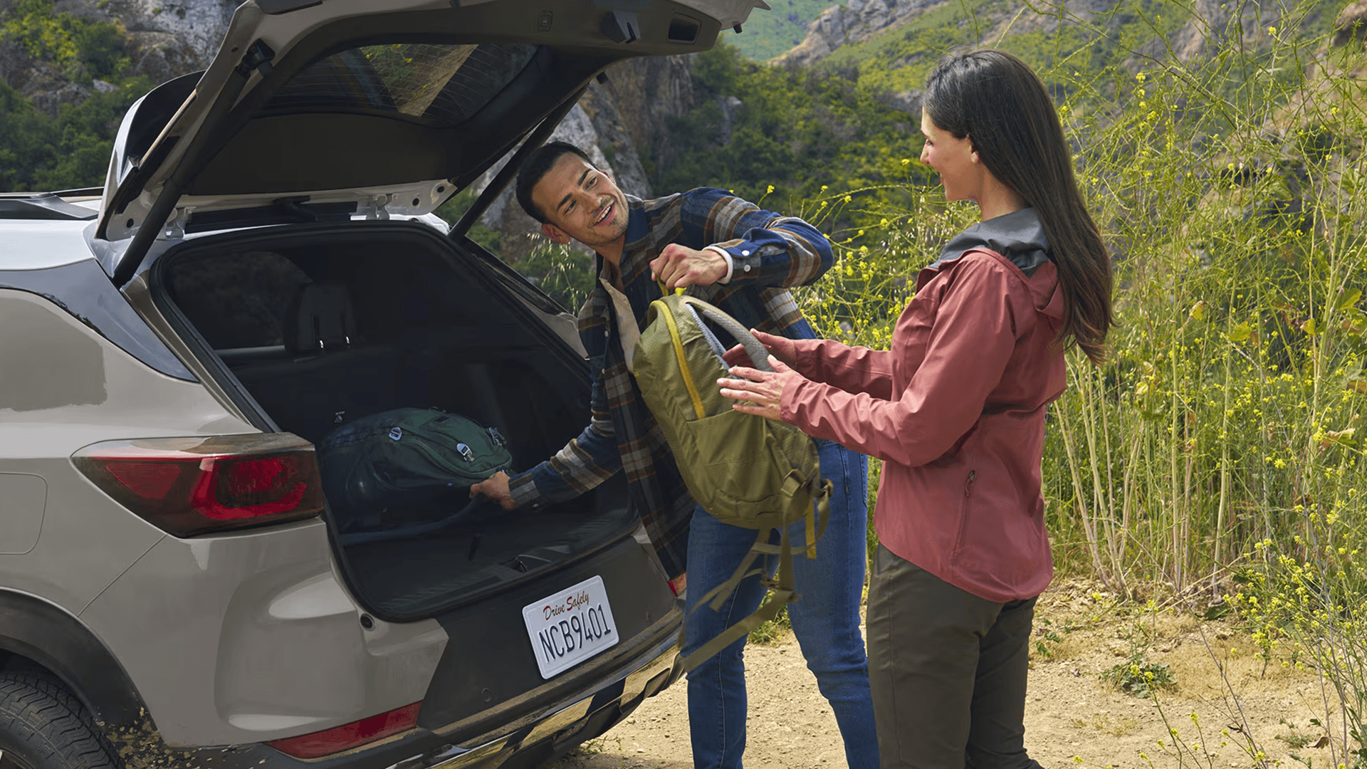 Two people load backpacks into the open rear cargo area of a light tan Chevy Trailblazer parked on a dirt trail near rocky hills.