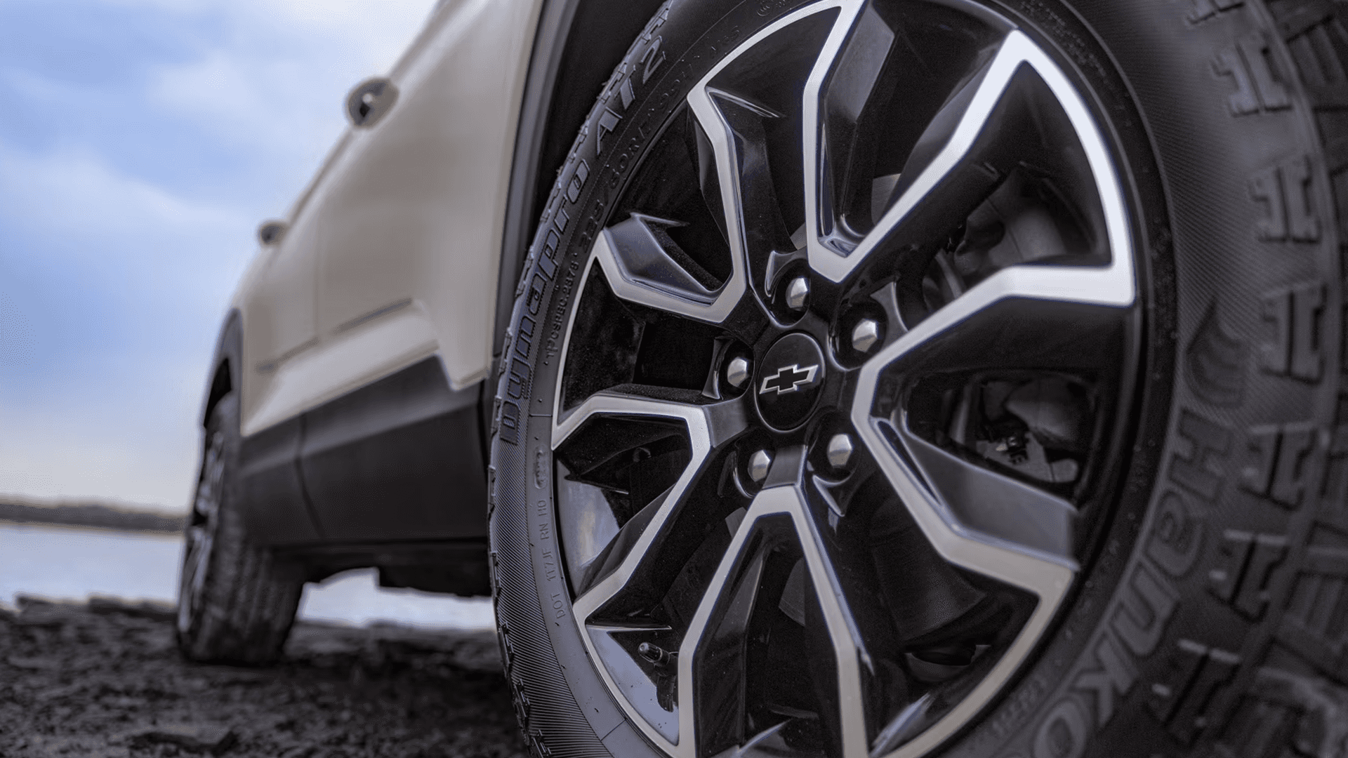 Close up of a black and silver Chevy alloy wheel with a Chevrolet bowtie center cap and all season tire near a lakeside setting.